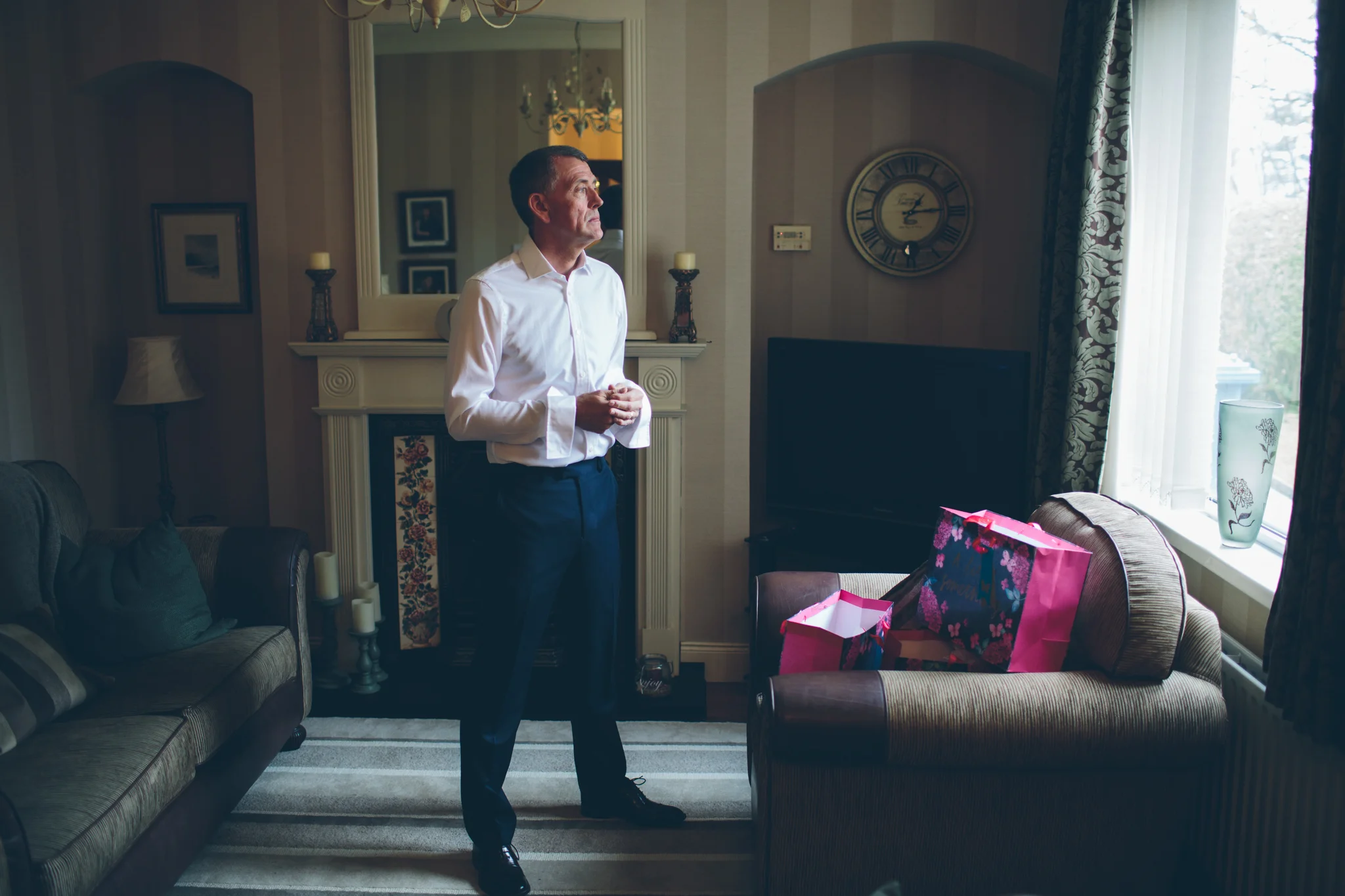 A man dressed in a white shirt and dark pants stands in a living room, looking out the window near a decorated fireplace with candles. There are gift bags on a nearby chair, and a large flat-screen TV is visible.