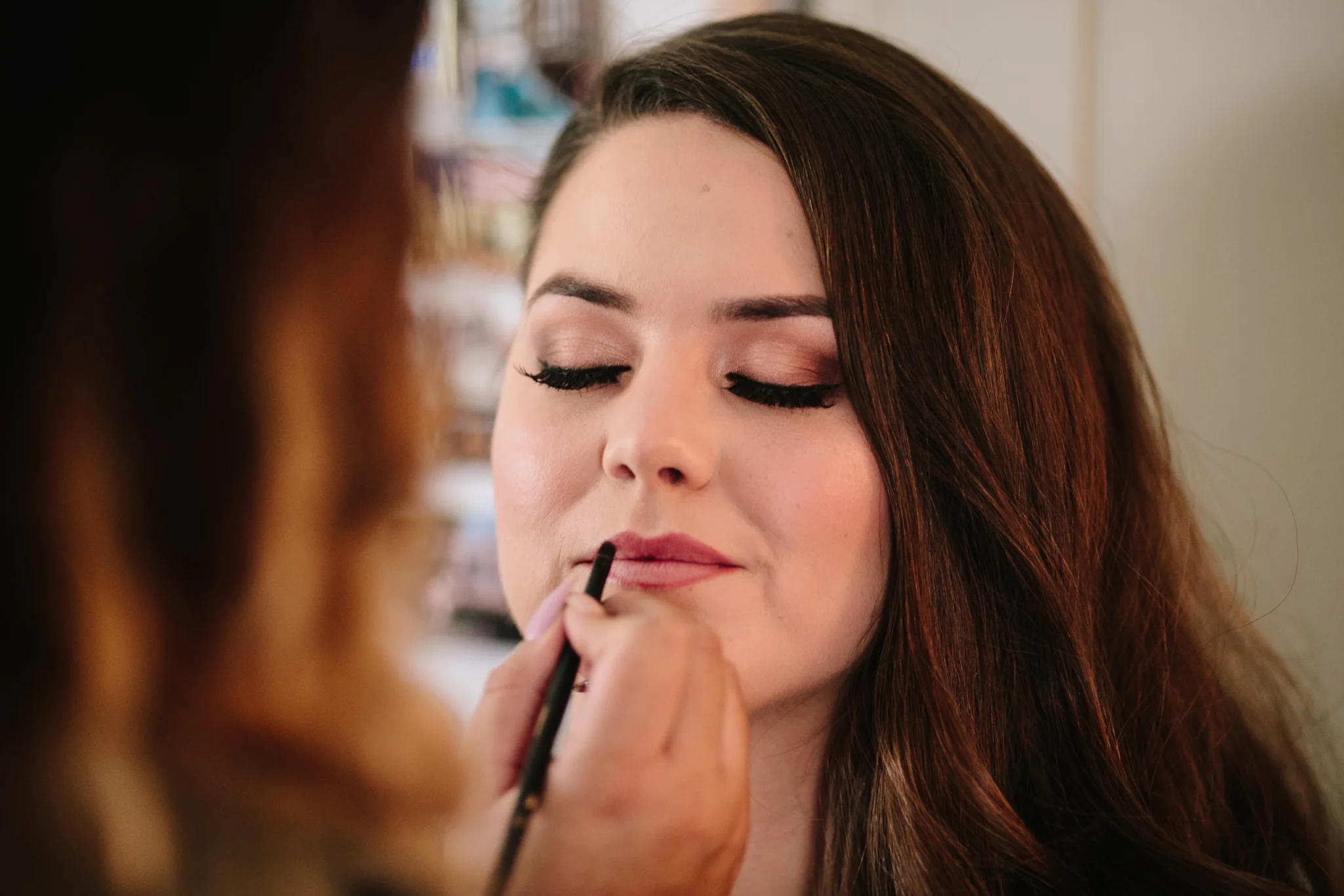 Close-up of a woman with closed eyes having lipstick applied by makeup artist in a beauty salon.