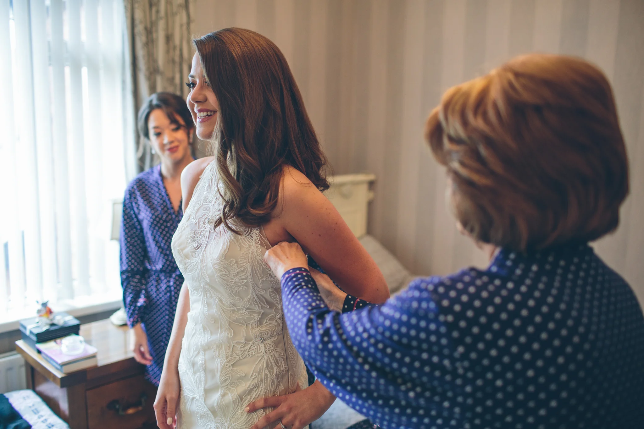 A bride is getting ready, assisted by women in a warmly lit room. One woman helps the bride by adjusting her dress, while another looks on smiling.