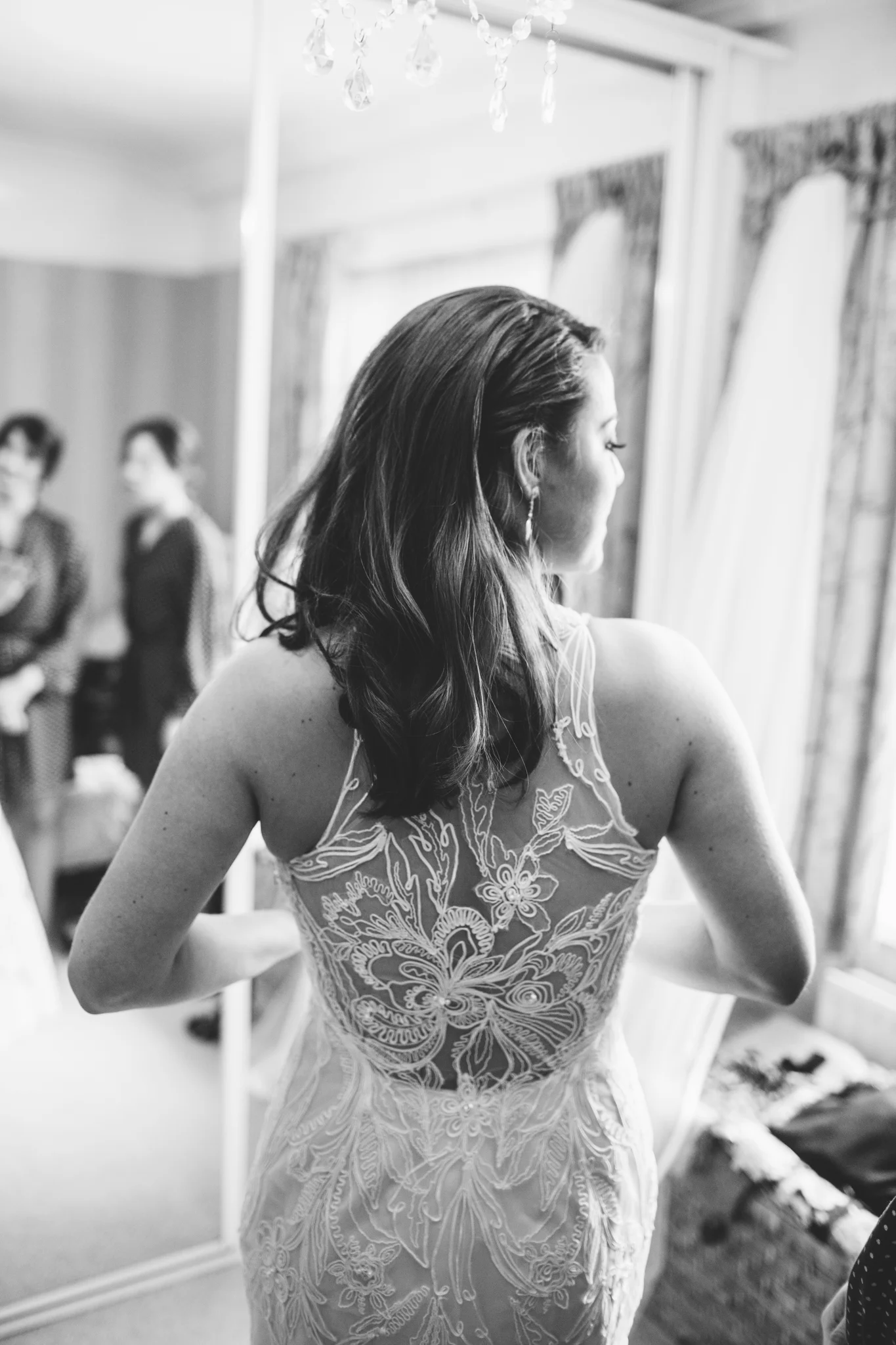 Black and white photo of a woman with long hair, wearing a lace dress, standing in front of a mirror, with three people in the background.