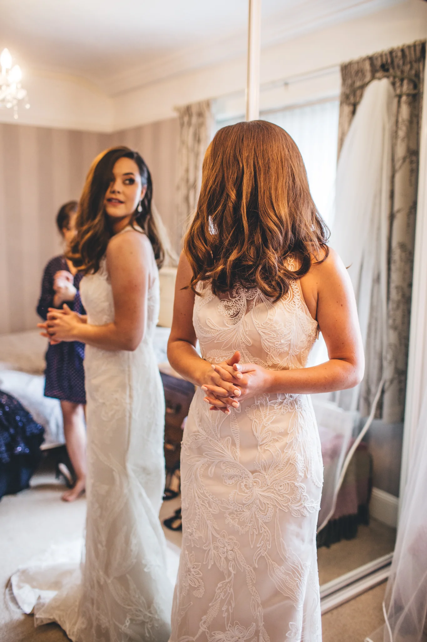 Two women in wedding dresses, one with red hair and the other with dark brown hair, standing in front of a mirror in a bedroom. Other people are visible in the background.