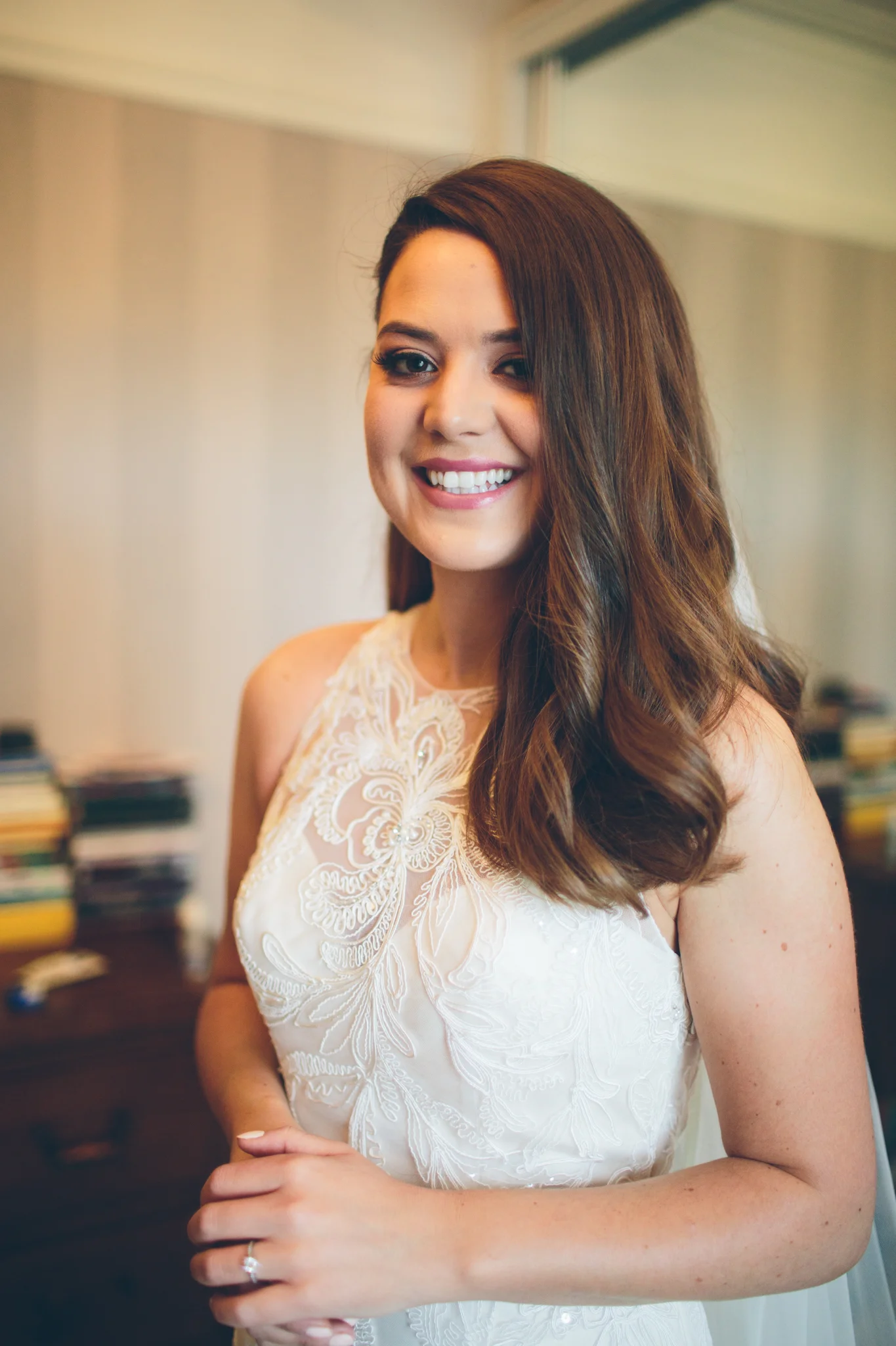 A woman with long wavy brown hair wearing a white lace dress, smiling indoors, with a blurred background of books and furniture.
