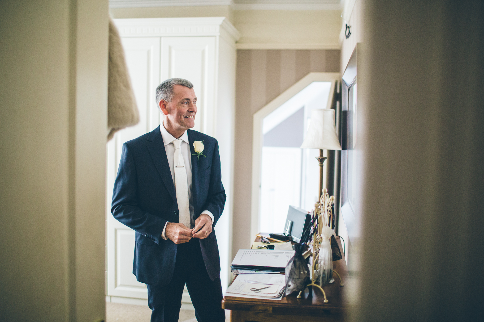 A man in a dark suit with a white shirt and tie, wearing a white flower boutonniere, standing inside a well-lit room, smiling and looking to his left.