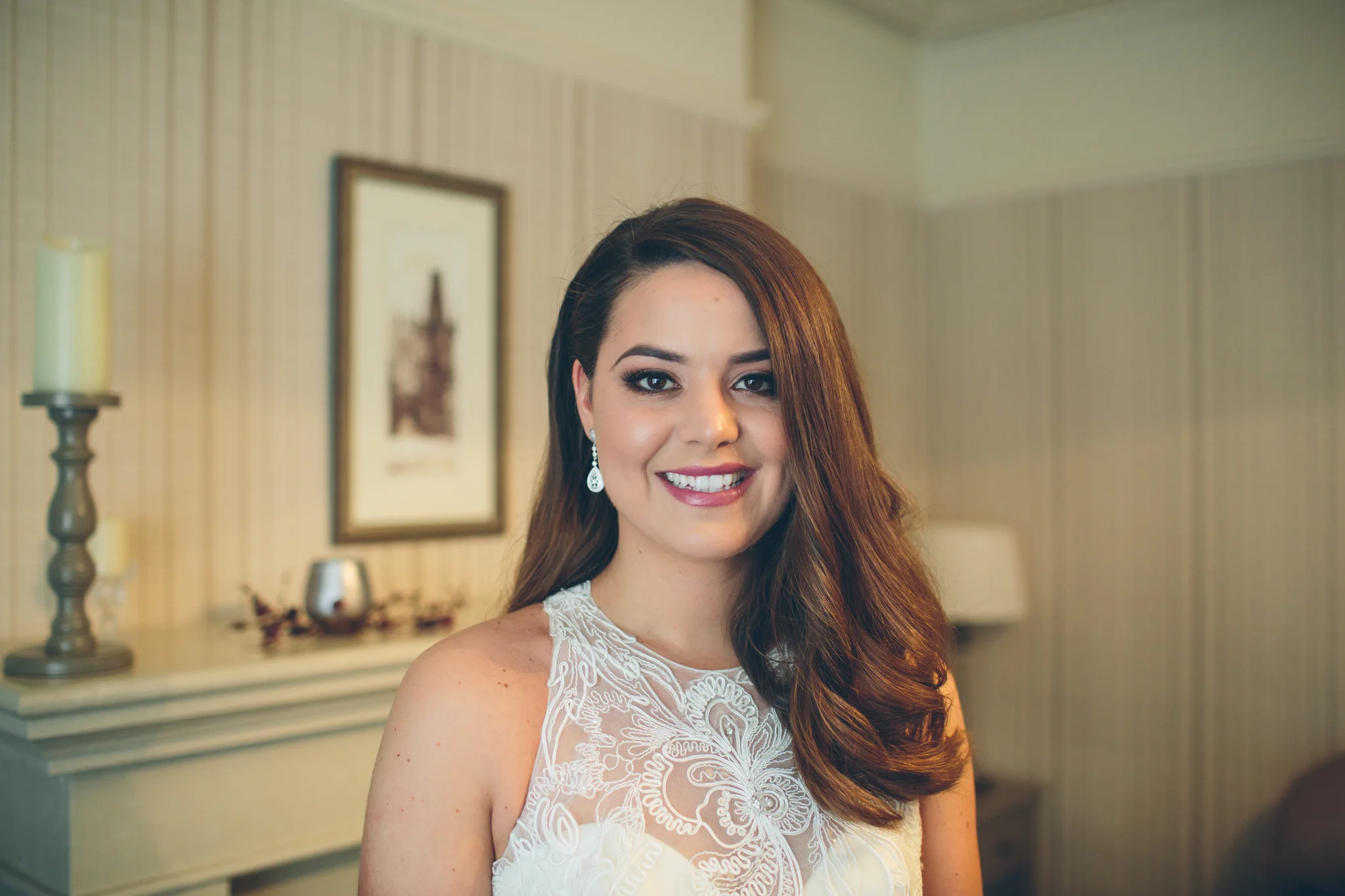 A woman with long brown hair styled in loose waves, wearing a white lace dress and teardrop earrings, smiling in a warmly lit room with framed artwork on the wall and a mantle with candles in the background.