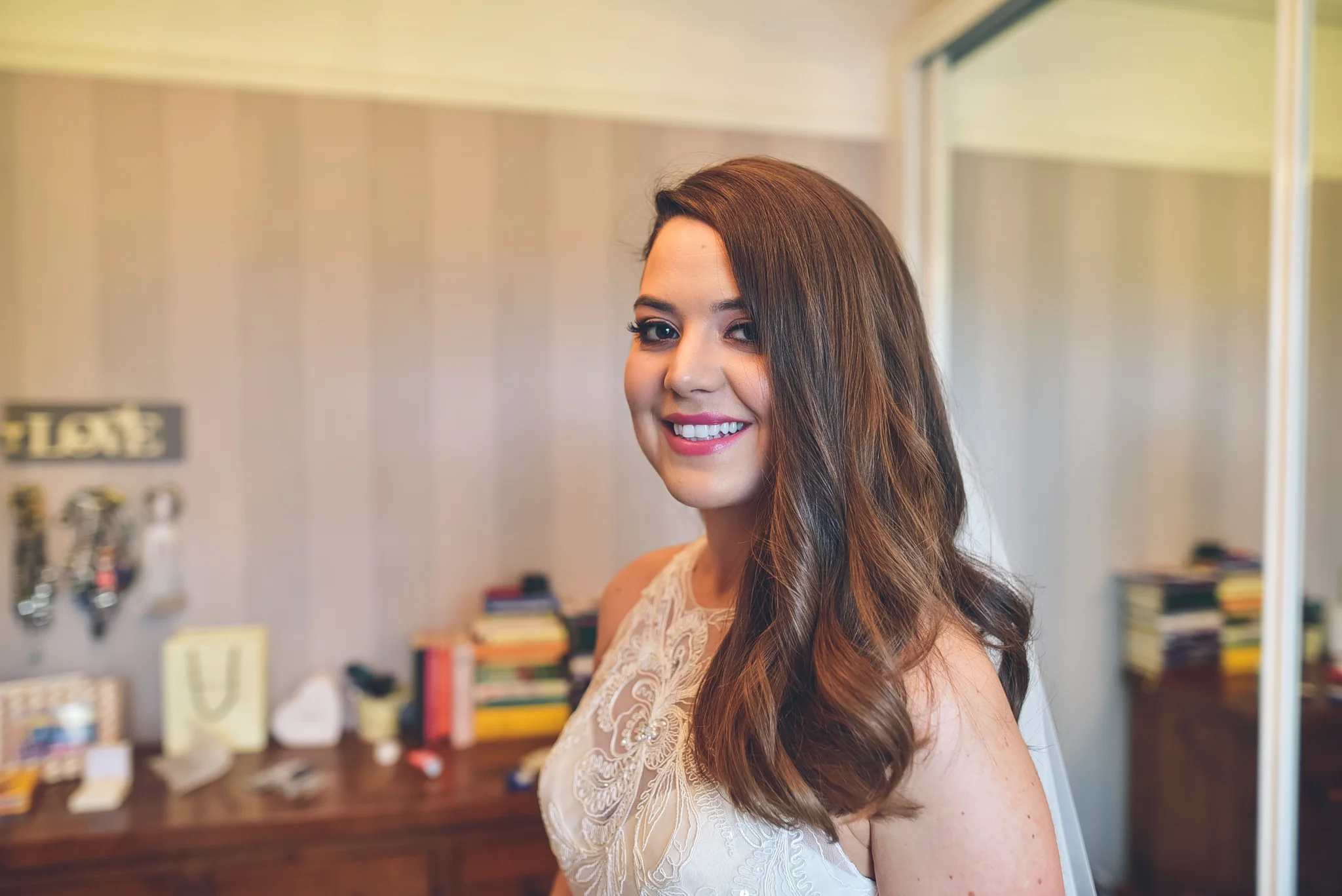 A woman with long, wavy brown hair and a bright smile, wearing a white lace dress, standing in a room with beige walls, a wooden dresser, and a wall with hooks and books in the background.