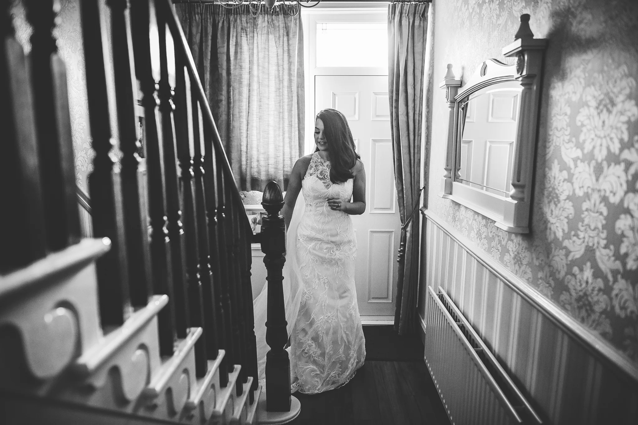 A woman in a white lace wedding dress standing in a hallway near a staircase, looking down and smiling. There is a window with curtains and a mirror on the wall.