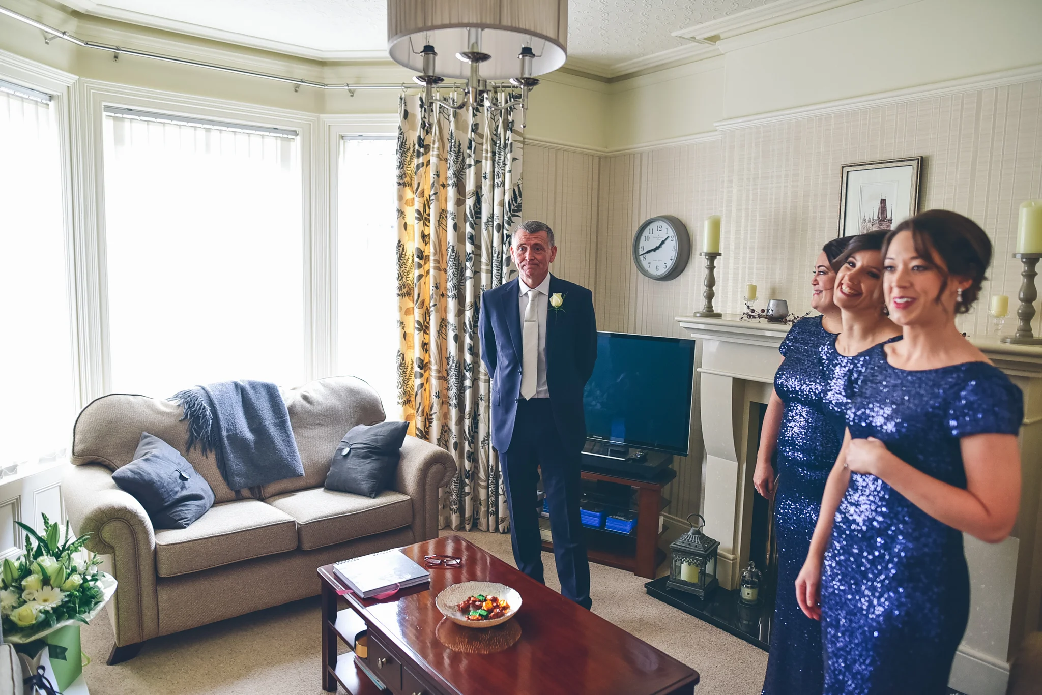 Three women and one man standing in a living room, with the women dressed in blue sequin dresses and the man in a suit. The man has a white rose on his lapel. There is a sofa with navy blue pillows and a blanket, a wooden coffee table with a bowl of 