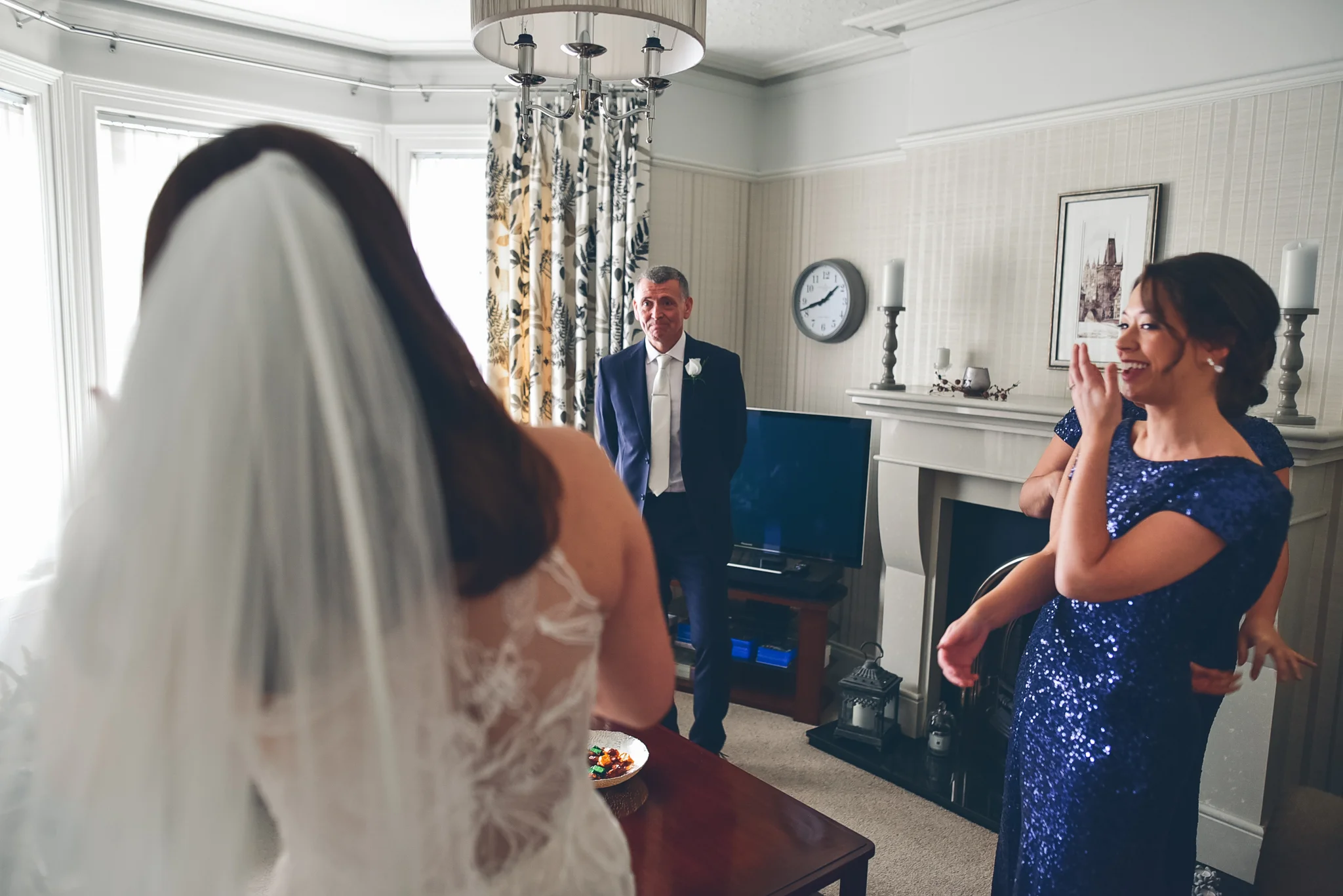 A woman in a wedding dress and veil is in the foreground with her back to the camera, facing a man in a dark suit and tie and a woman in a blue sequin dress, who are both smiling. The scene takes place inside a living room with a fireplace, a clock, 