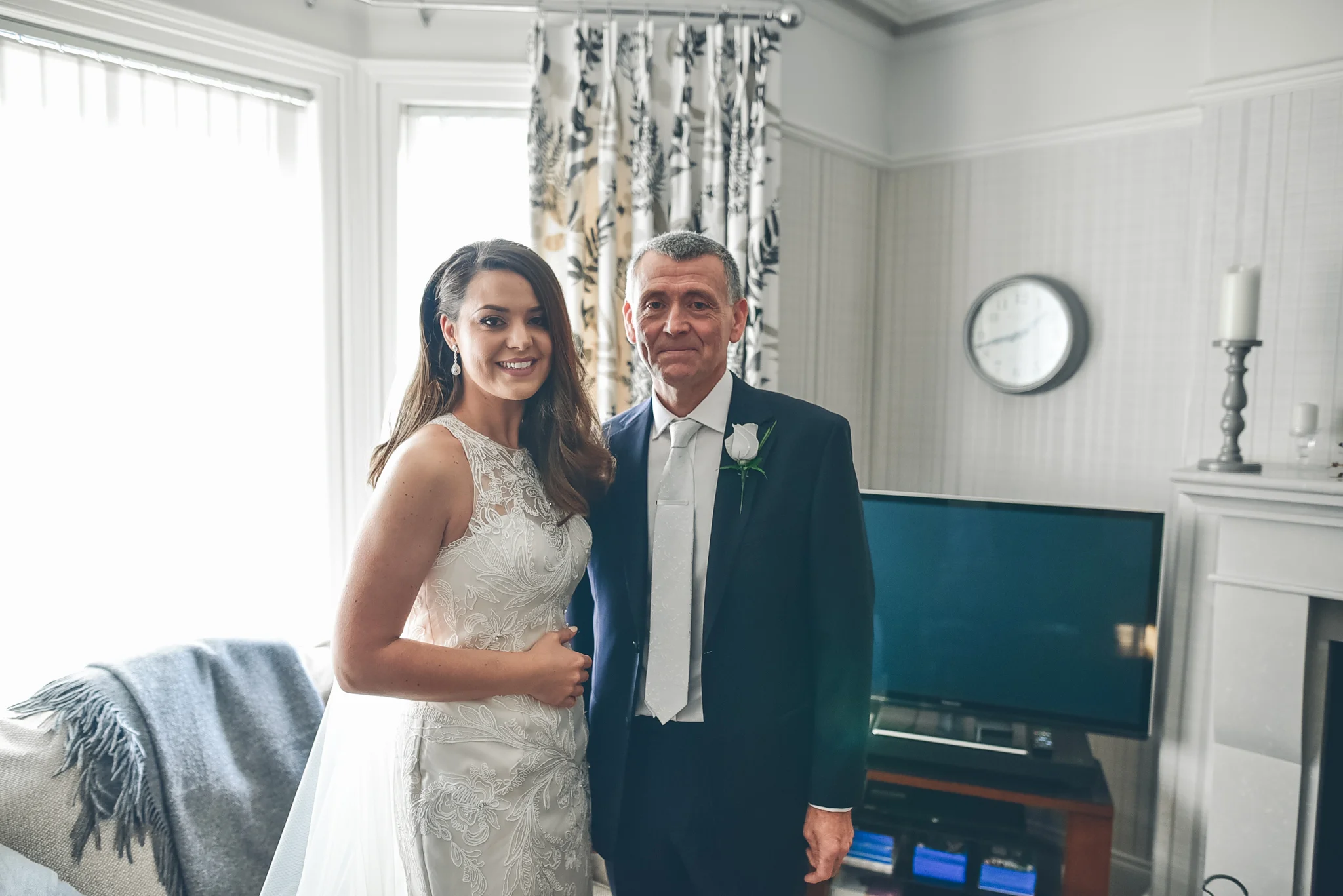A young woman in a white wedding dress standing next to an older man in a suit with a white boutonniere, inside a living room with a television, clock, and curtains.