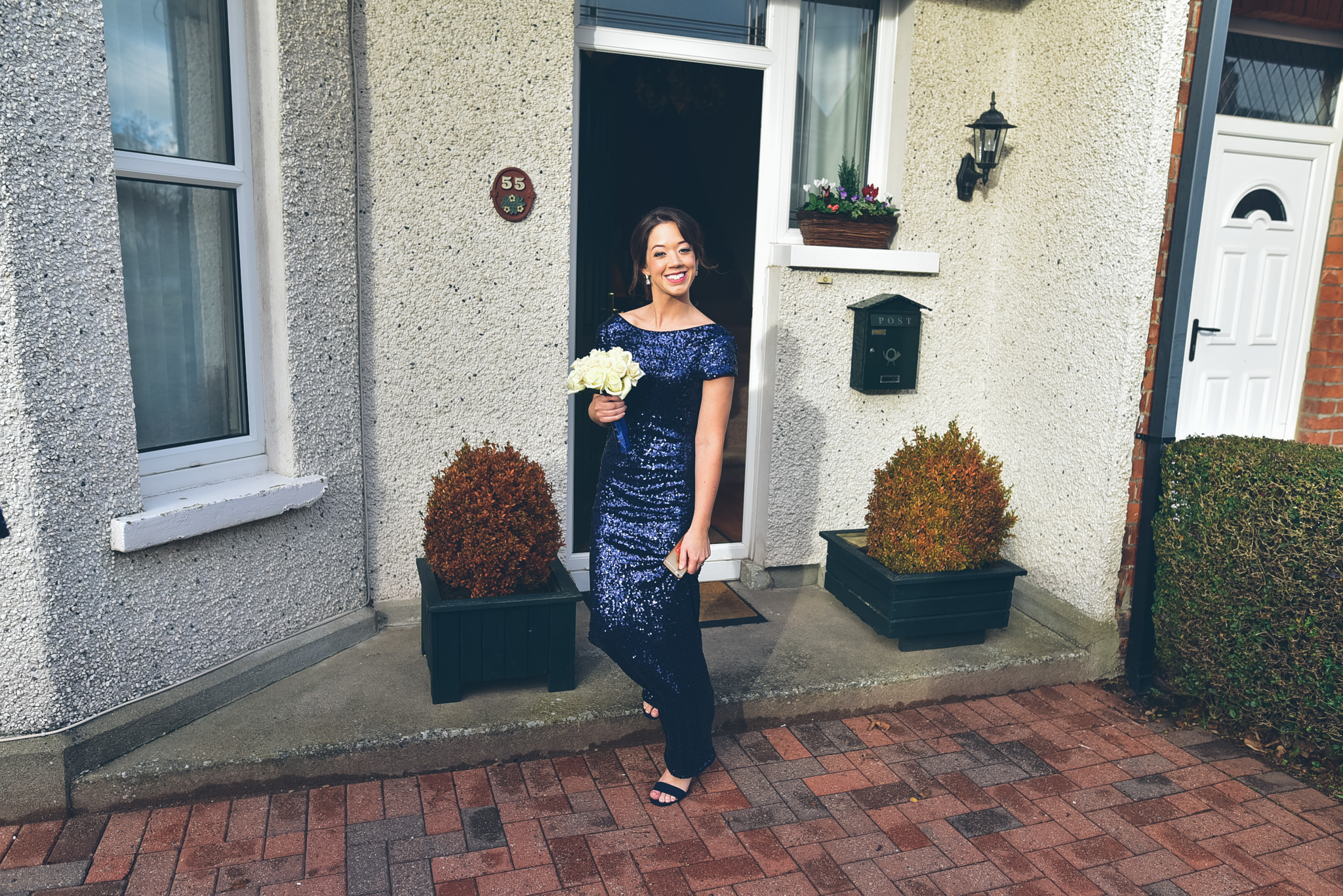 Woman in a blue sequin dress holding a bouquet of white roses standing in front of a house exterior with a white door, window, potted plants, and bushes.