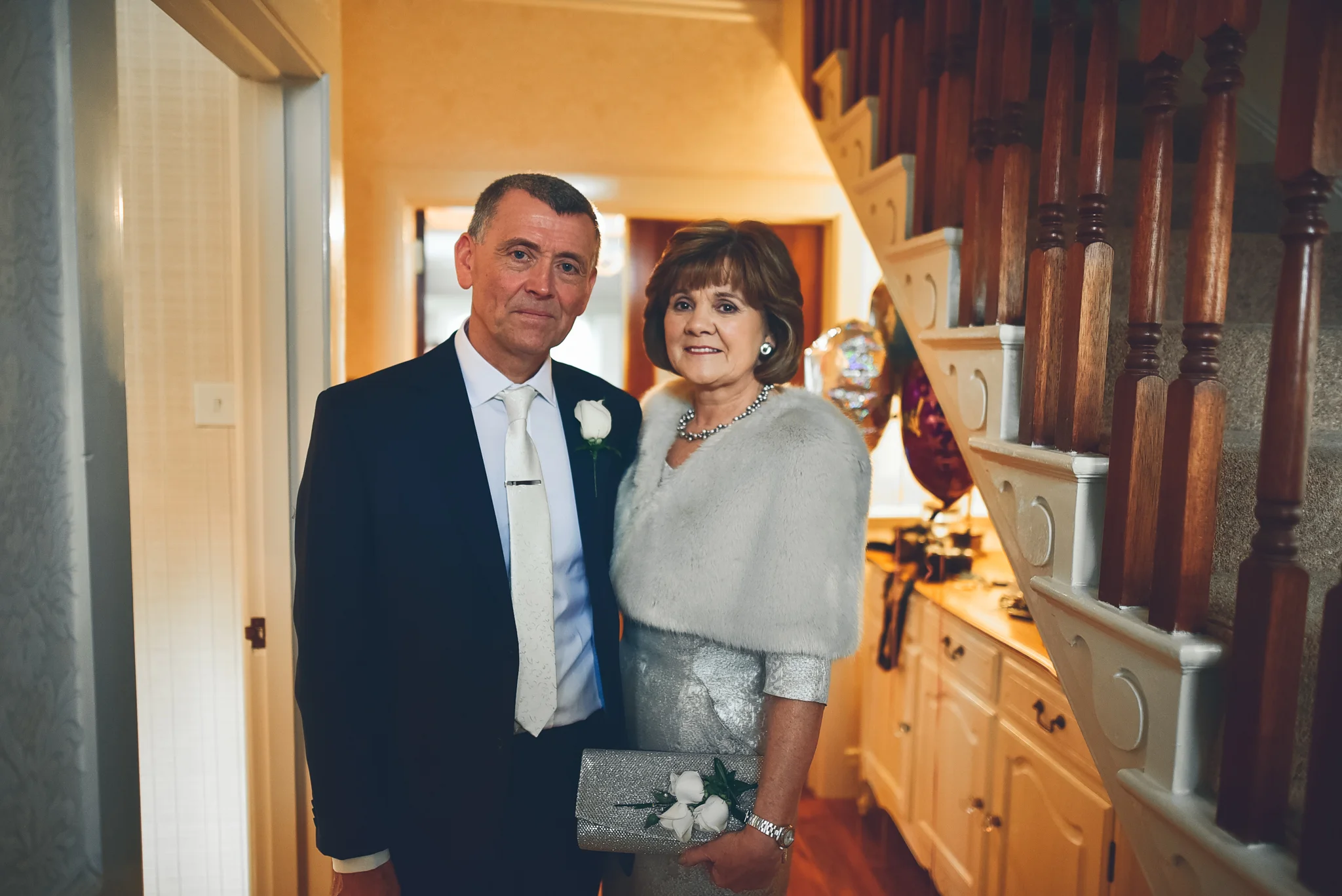 A middle-aged man and woman dressed formally, standing inside a house. The man wears a dark suit with a white shirt, white tie, and a white flower boutonniere. The woman wears a sparkly dress with a light-colored shawl and holds a silver clutch purse