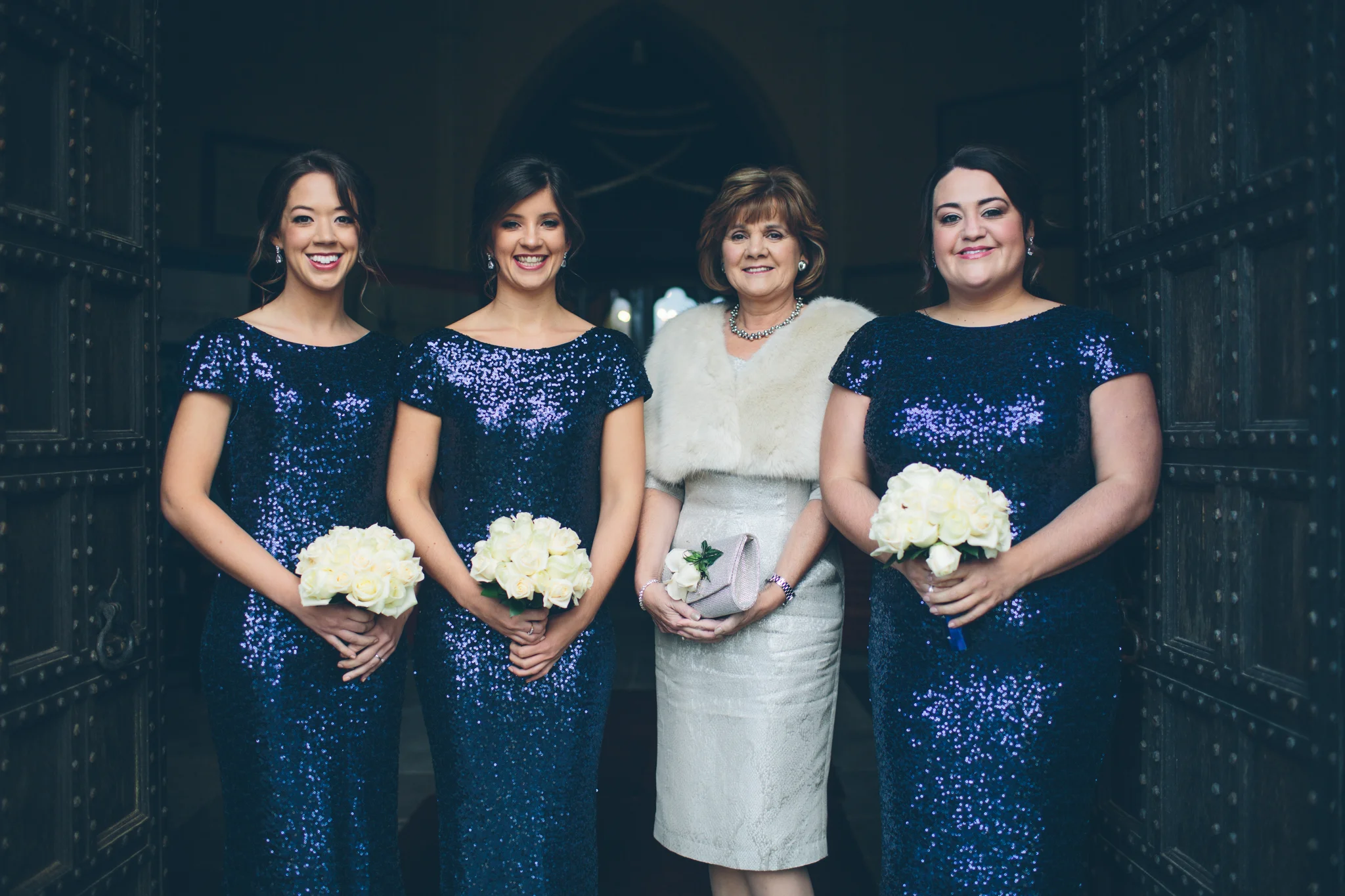 Four women in formal dresses posing together at a wedding or special event, holding bouquets of white roses, with a dark background and large wooden doors.