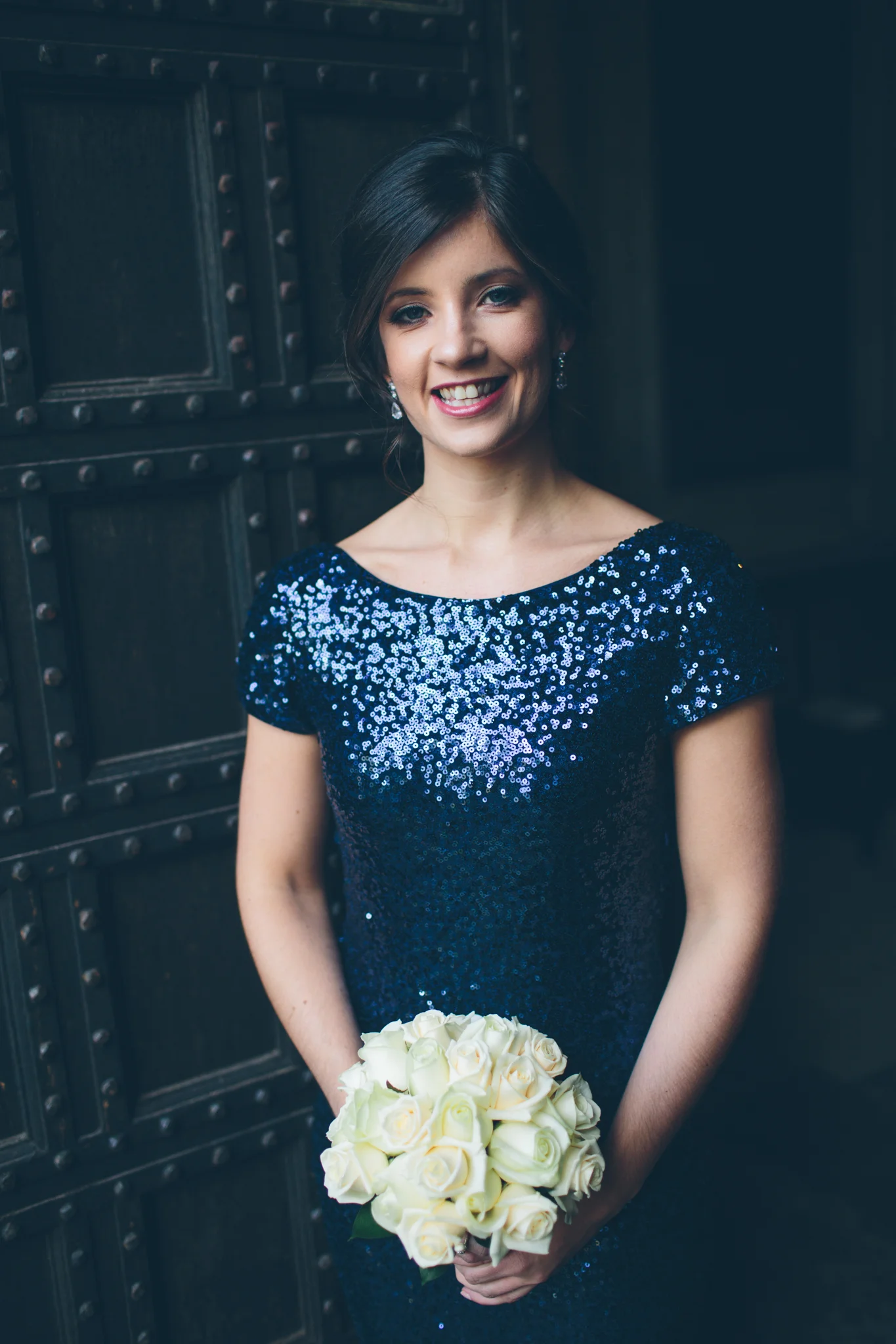 A woman in a dark blue sequined dress holding a bouquet of white roses, smiling in front of a dark industrial background.
