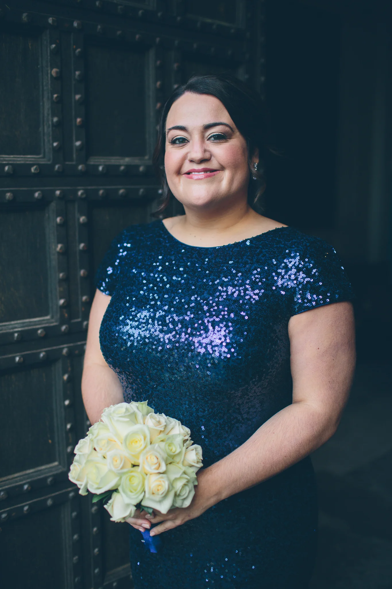 A woman in a dark blue sequin dress holding a bouquet of white roses, standing in front of a dark, textured background.