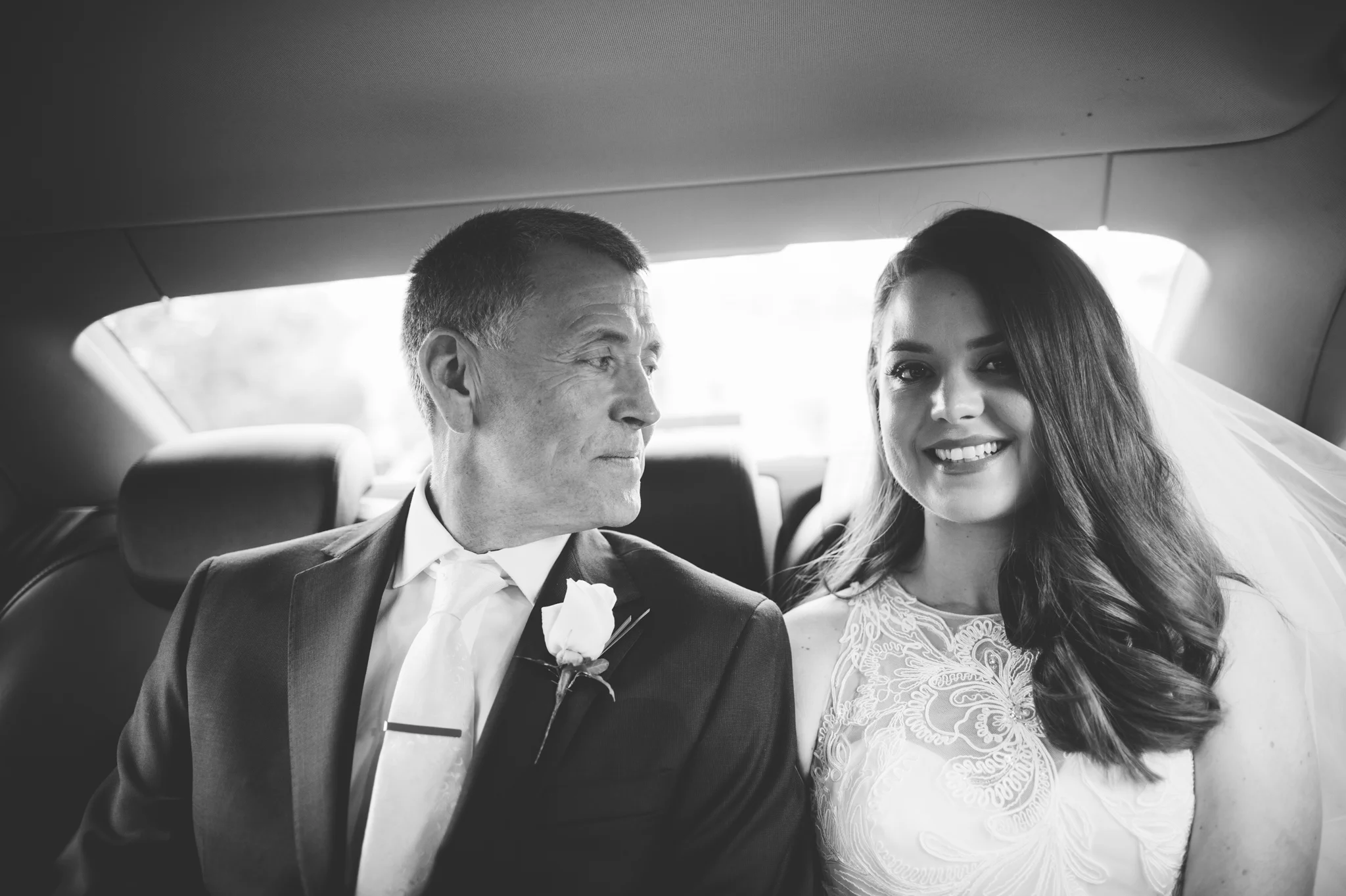 A bride with long, wavy hair smiling, sitting beside an older man in a tuxedo with a white flower boutonniere, inside a car with a window in the background.