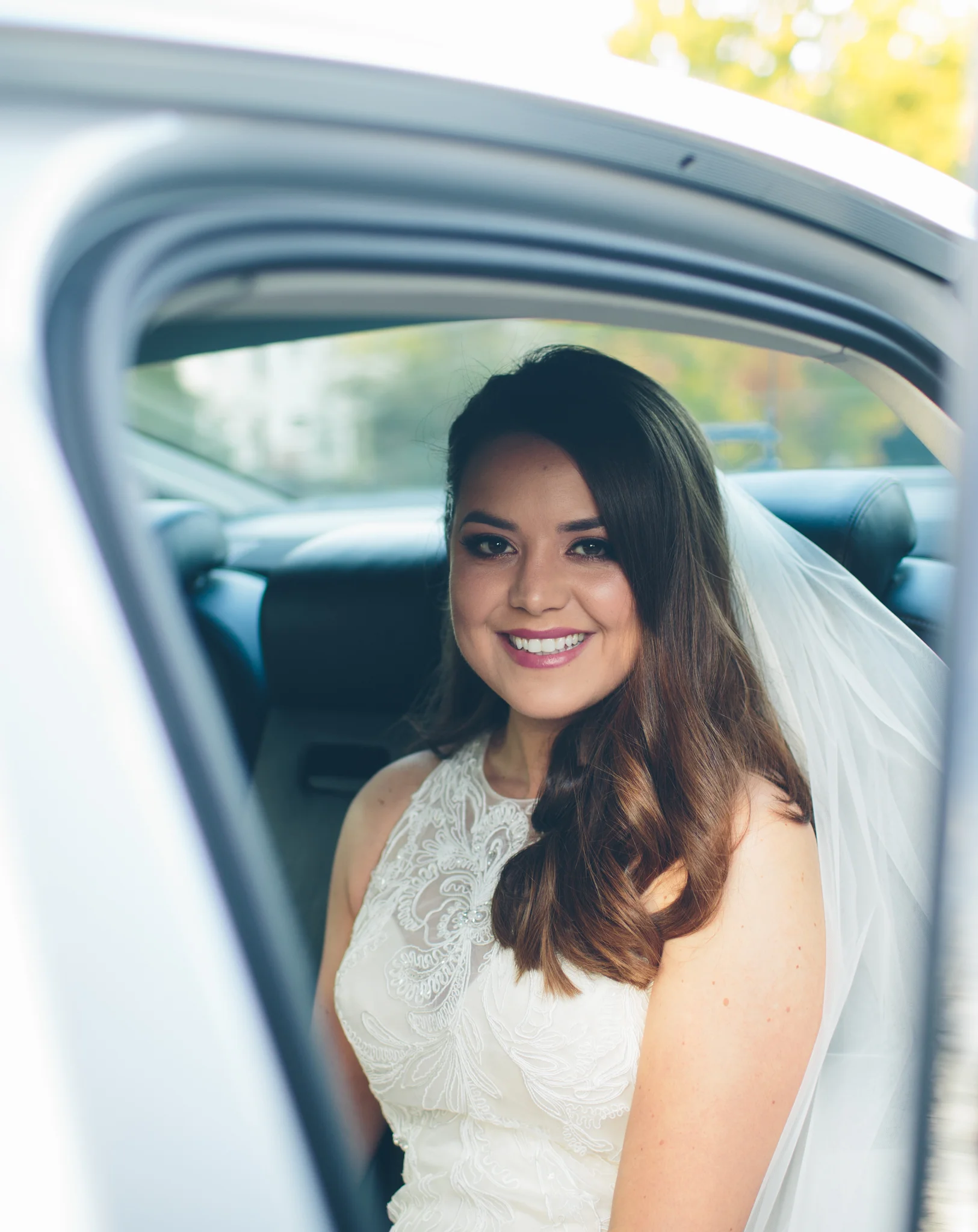 A smiling bride in a white lace wedding dress and veil sitting in the backseat of a car, looking out the window.