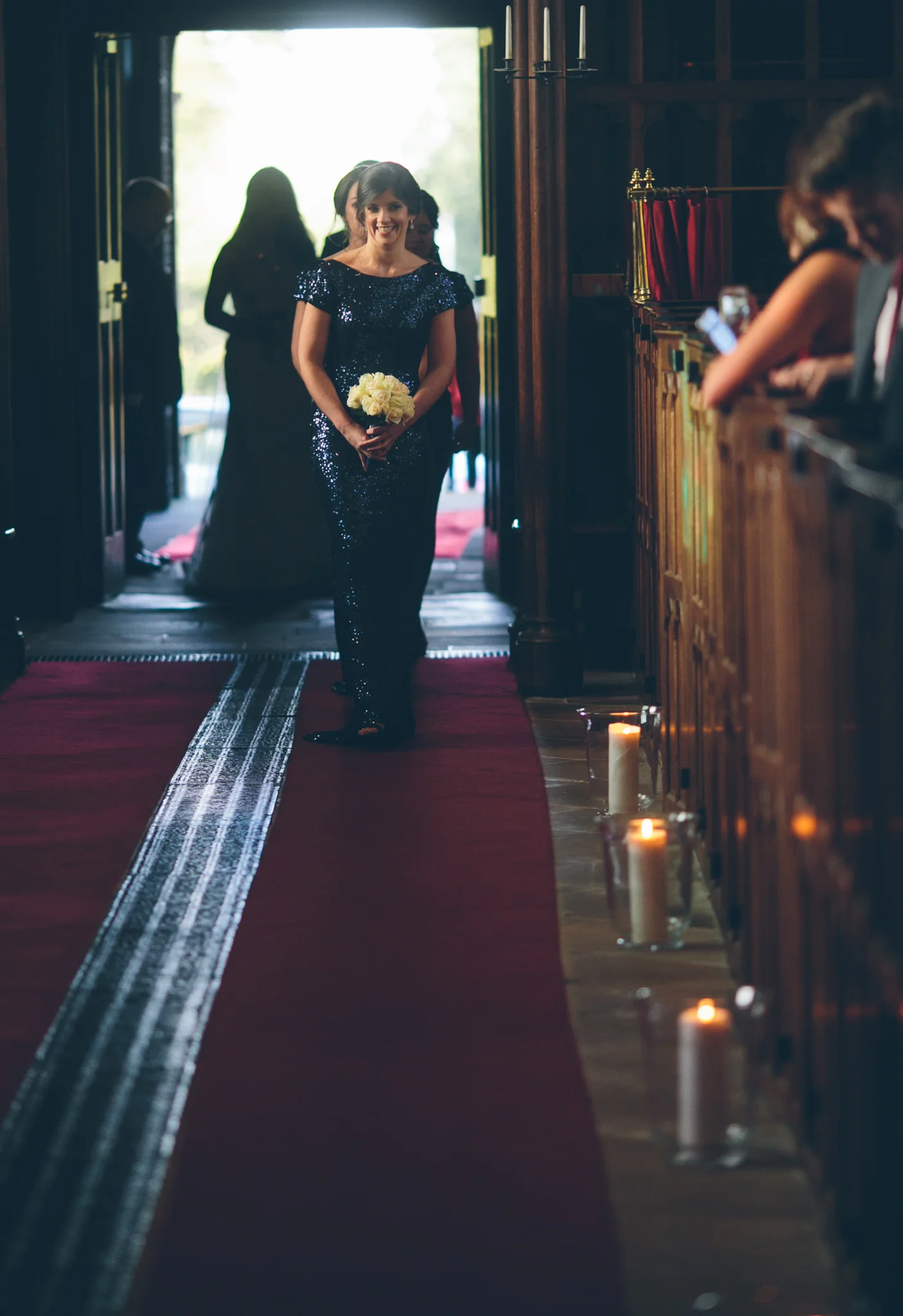 A woman in a dark blue, sequined dress holding a bouquet of white roses walking down an aisle in a church. Several candles in glass holders line the side of the aisle. People are seated along the right, and a bright open door is in the background wit