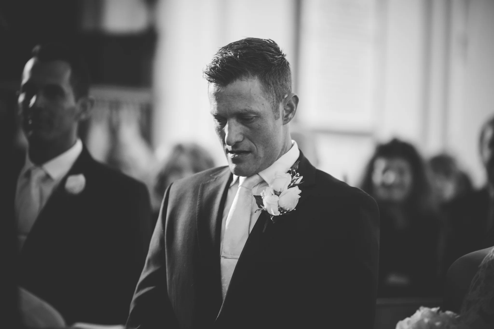 Black and white photo of a groom during a wedding ceremony, standing with eyes closed, wearing a suit with a white boutonniere on his lapel, in a church or similar setting with blurred attendees in the background.