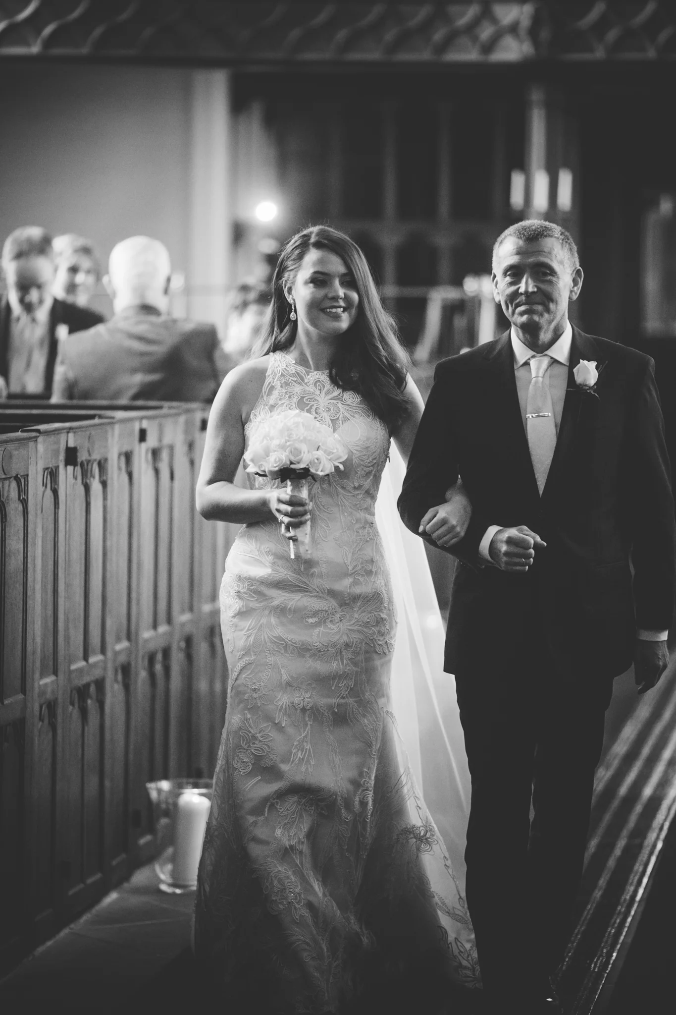 A bride walking down the aisle arm-in-arm with her father at a wedding ceremony, holding a bouquet of roses, inside a church.