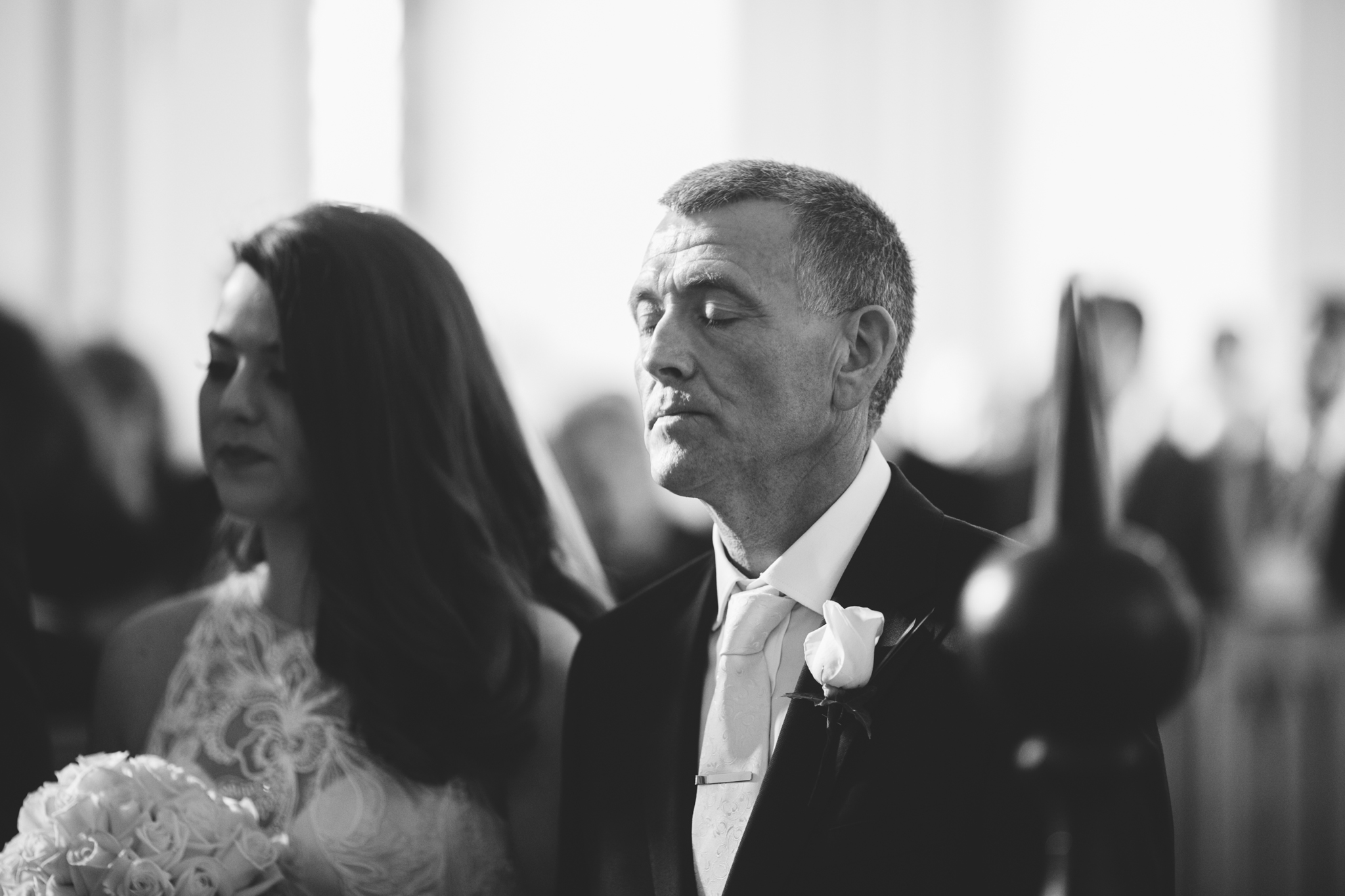 Black and white photo of a man and woman at a wedding ceremony, both with eyes closed, sitting side by side. The woman holds a bouquet of white roses, and the man is dressed in formal wear with a white boutonniere