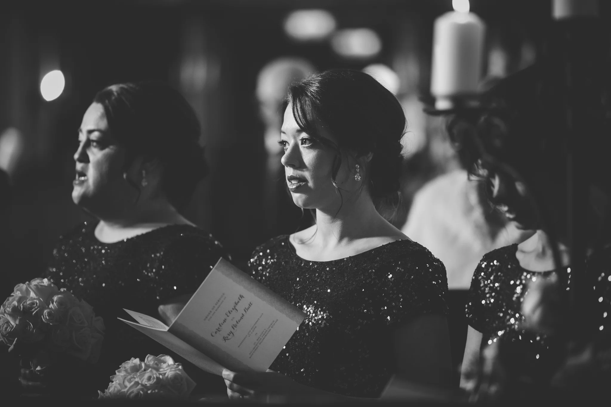 Black and white photo of women in elegant dresses, attending a ceremony or event, with one woman reading from a program and holding a bouquet of roses.