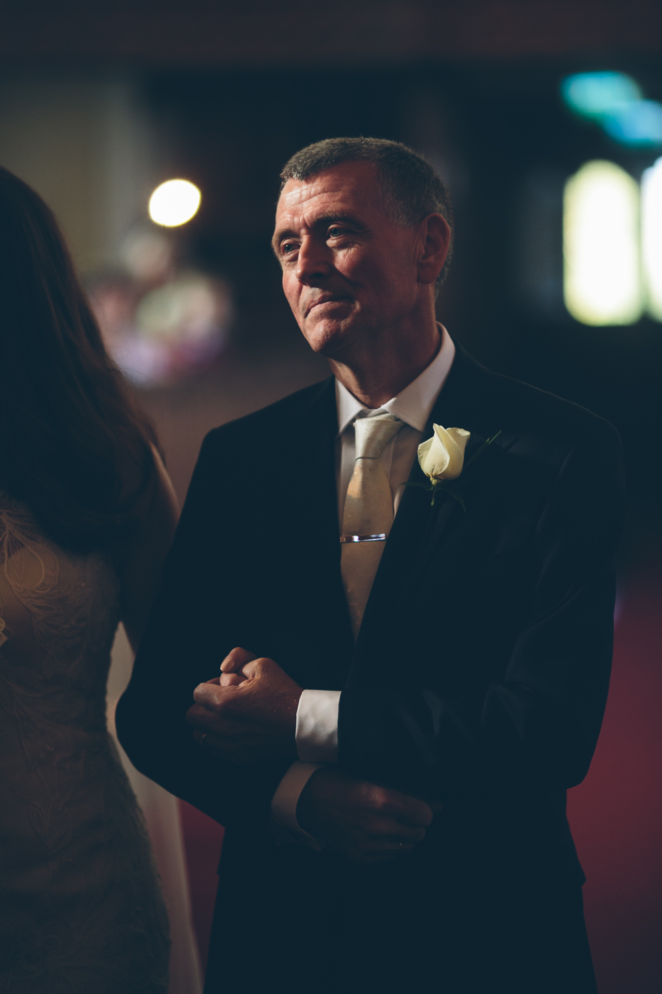 A man in a black suit with a white tie and a white rose boutonniere is standing with his arms crossed, looking contemplatively in a dimly lit setting with blurred background lights.