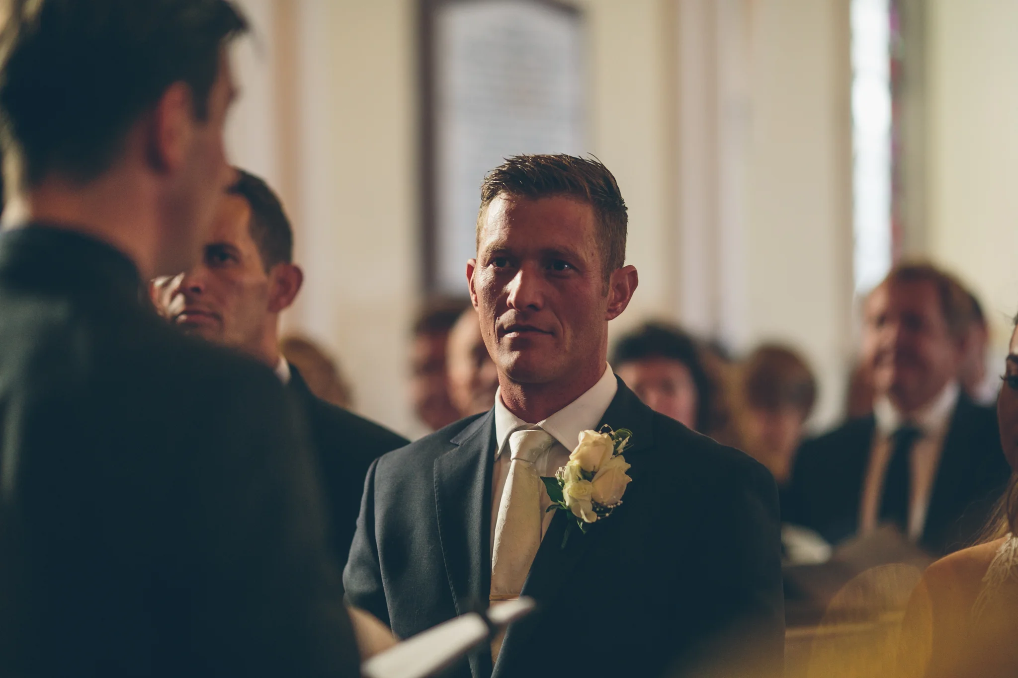A groom in a tuxedo with a white boutonniere stands in a church during a wedding ceremony, looking attentively at his vows.