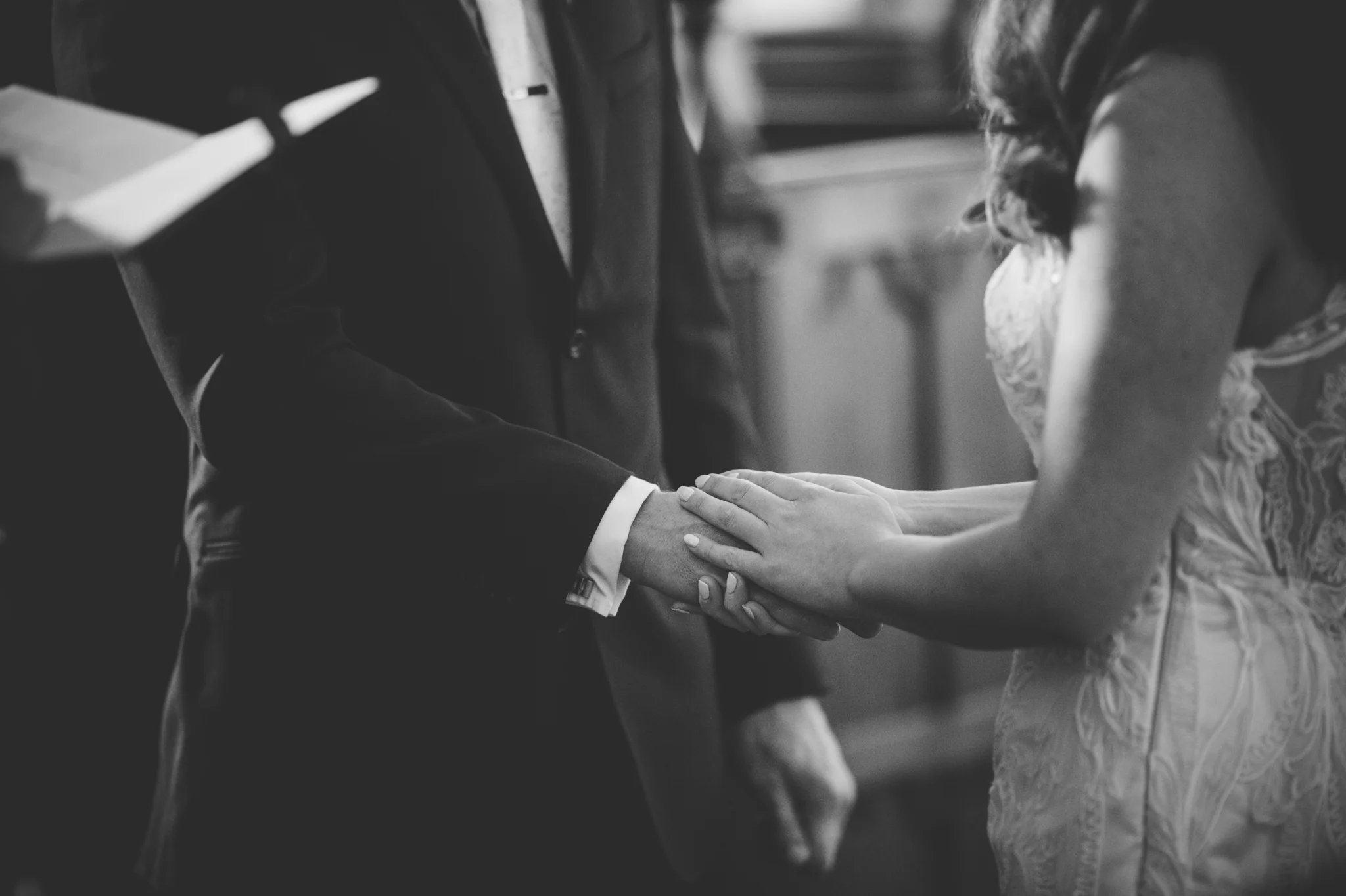 A black-and-white photograph of a wedding ceremony showing a bride and groom holding hands, with only their hands and parts of their bodies visible.