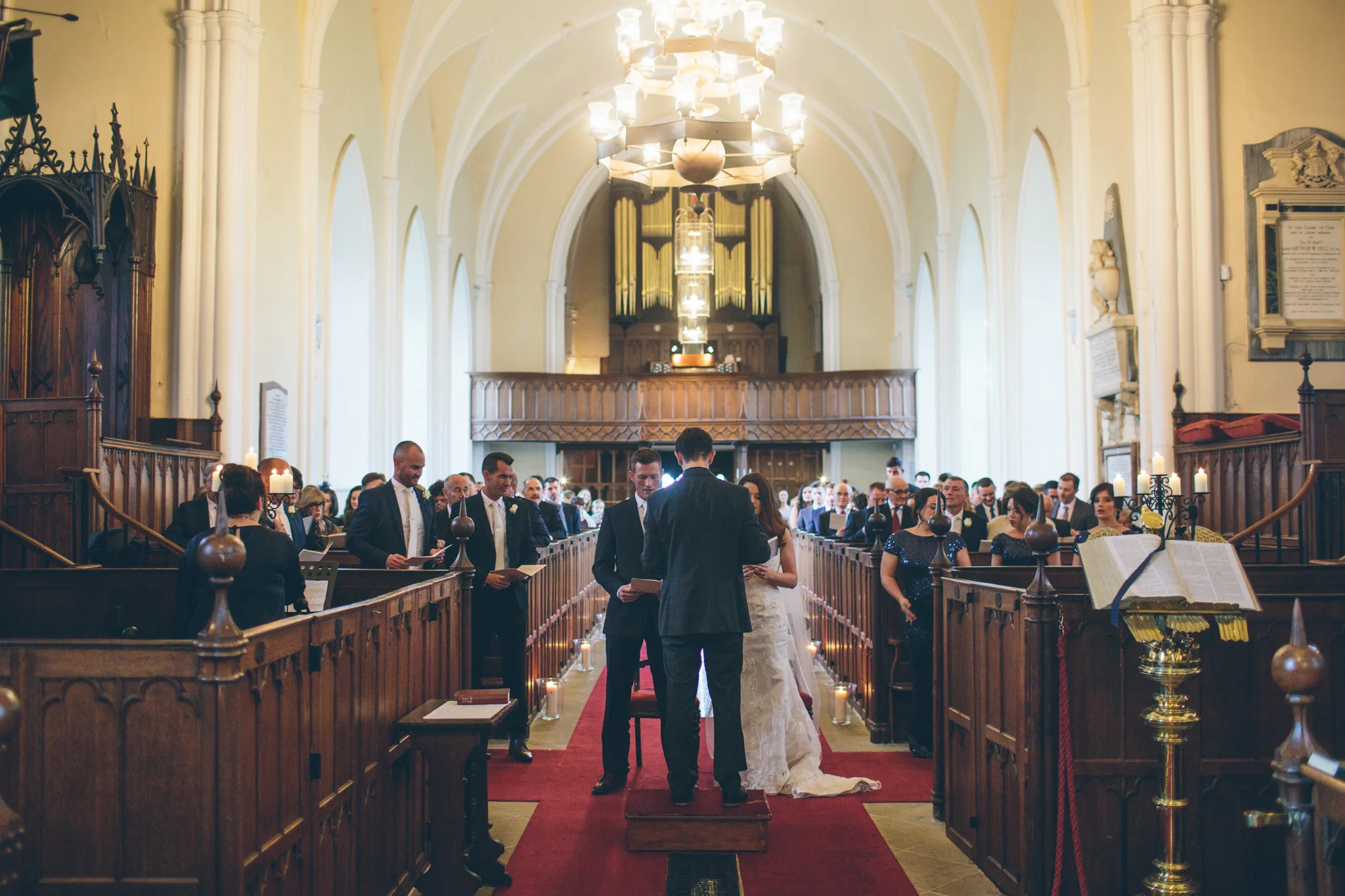 A wedding ceremony inside a church with the bride and groom standing at the altar, surrounded by family and friends seated in wooden pews.