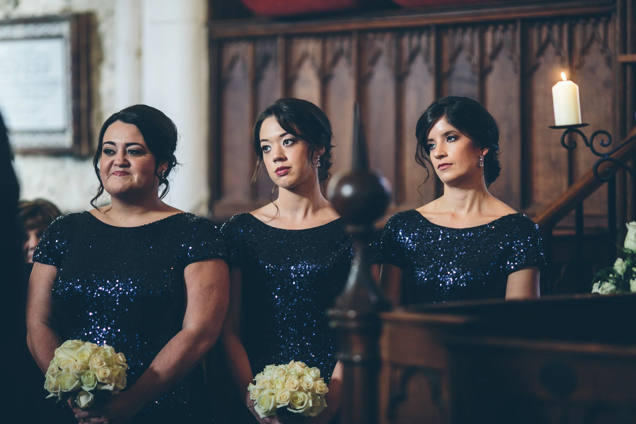 Three women at a wedding ceremony, wearing matching dark blue, sequined dresses, holding bouquets of white roses. They are standing in a church with wooden interior and lit candles.