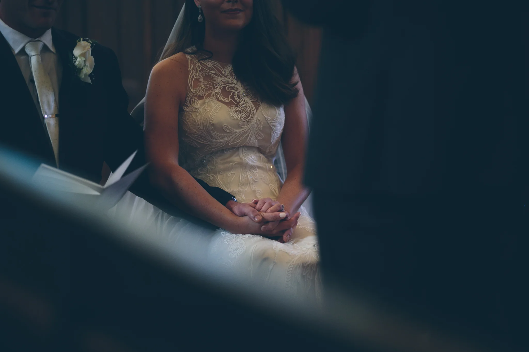 A bride and groom holding hands during a wedding ceremony, with the bride wearing a cream-colored lace dress and the groom in a dark suit with a boutonniere.