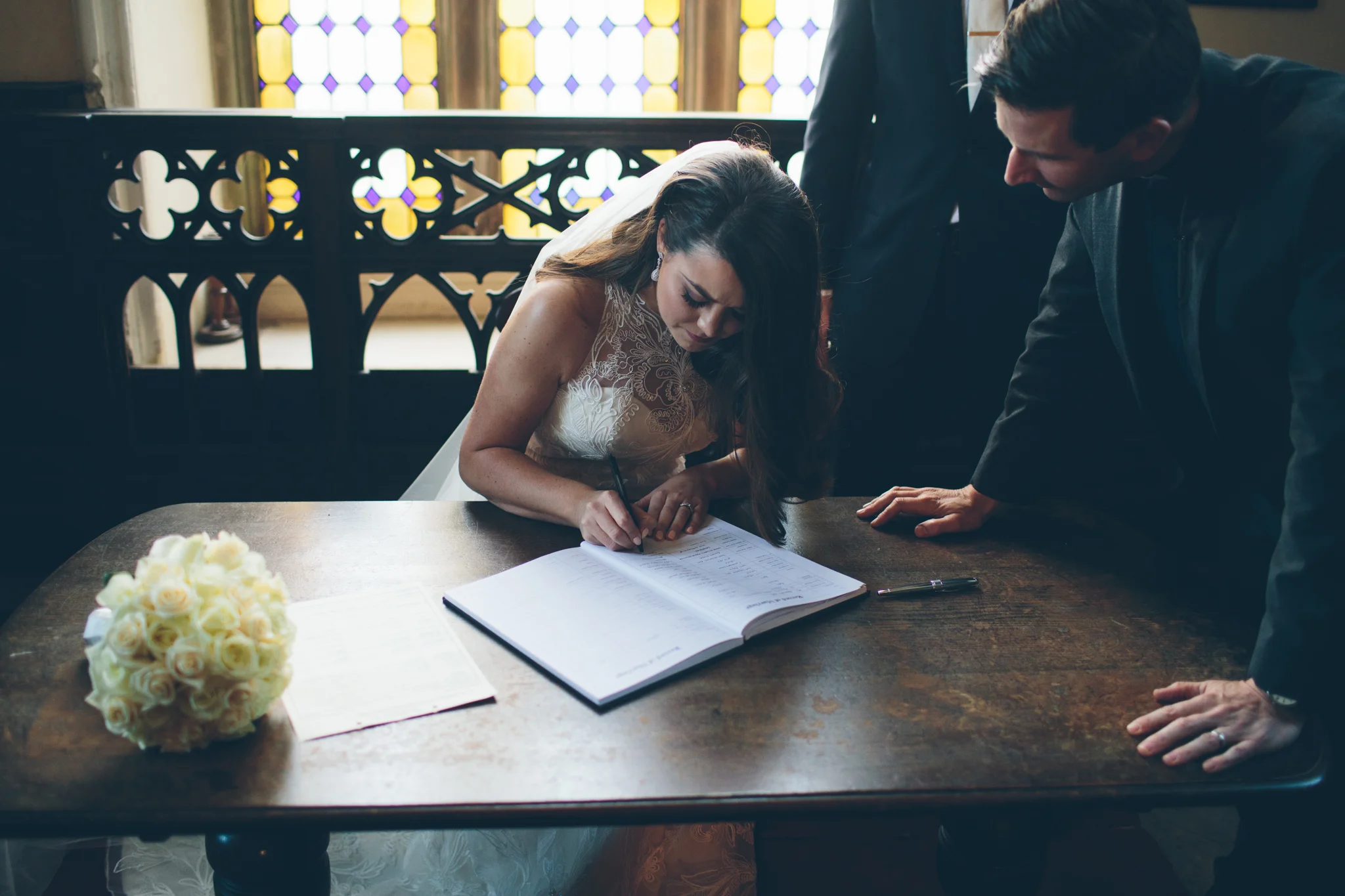 A bride signing a wedding register at a table during a wedding ceremony, with a bouquet of white roses on the table and a person leaning over to witness the signing.