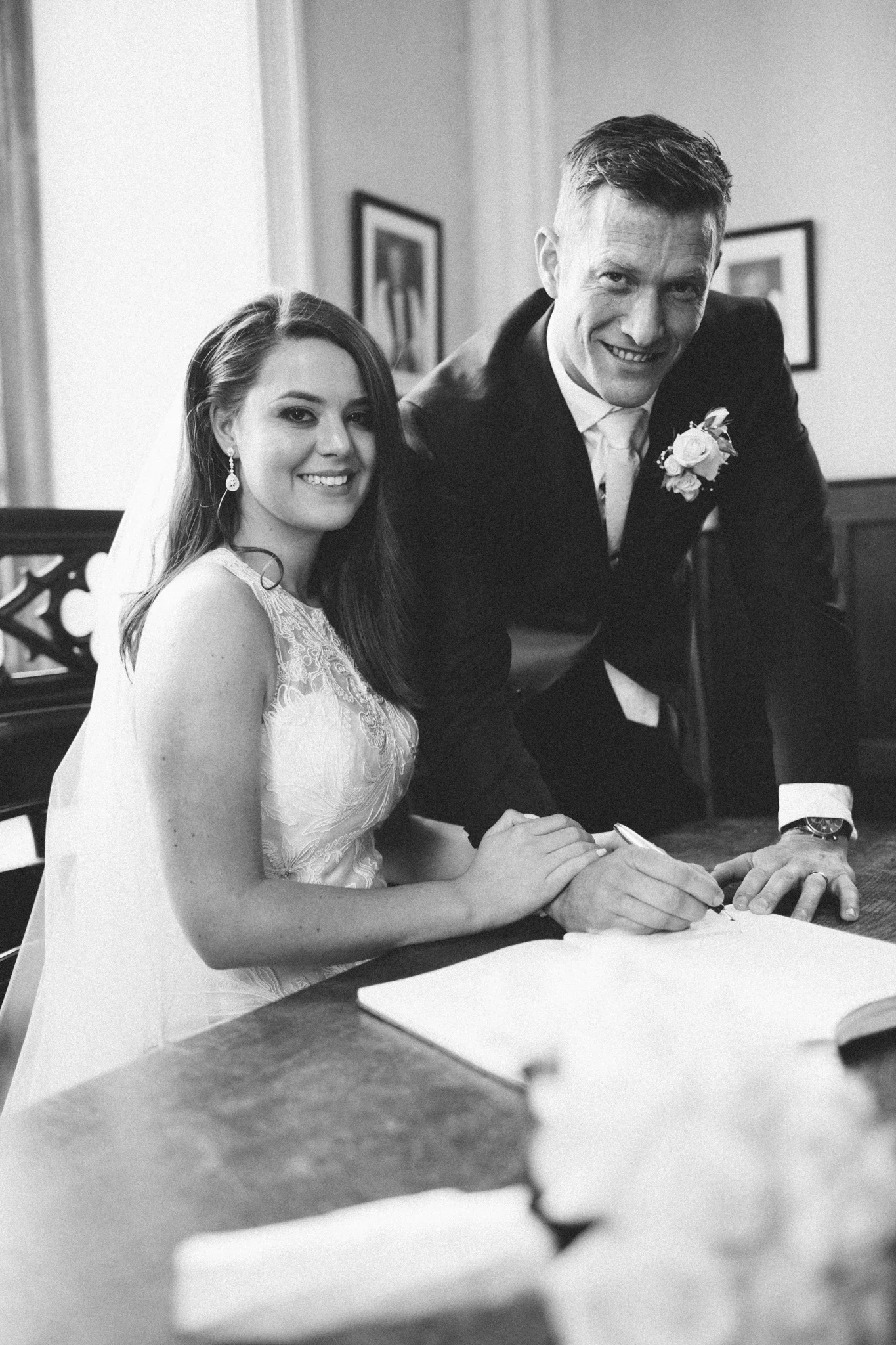 A black and white photo of a bride and groom signing a marriage document, with the groom leaning over the table and smiling.