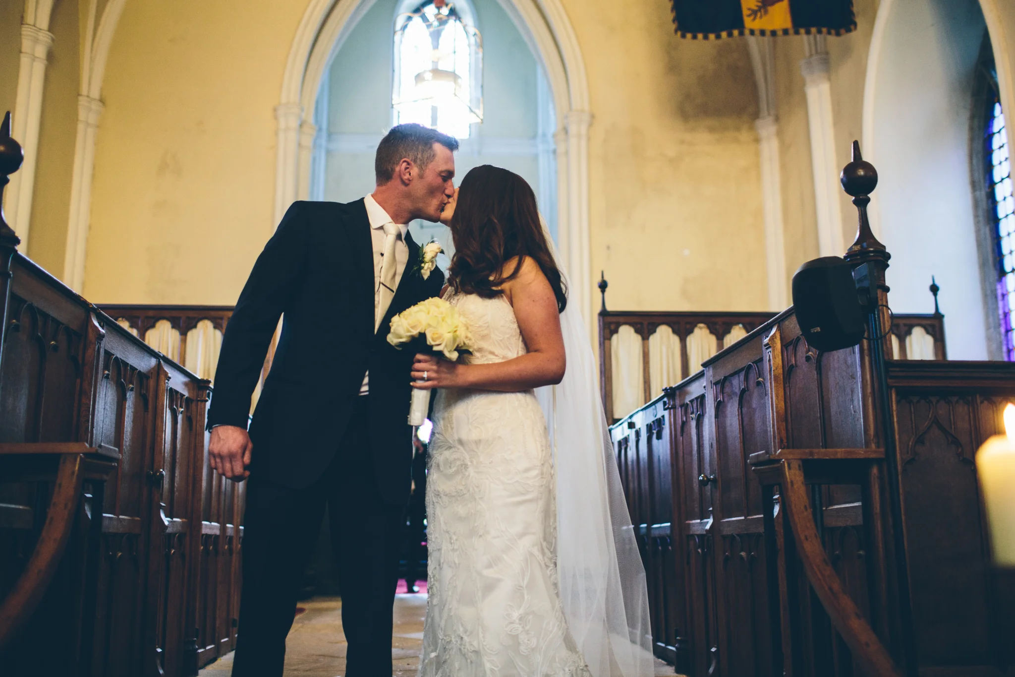 A bride and groom share a kiss inside a church during their wedding ceremony. The groom is in a black suit and the bride is in a white wedding dress holding a bouquet of white roses.