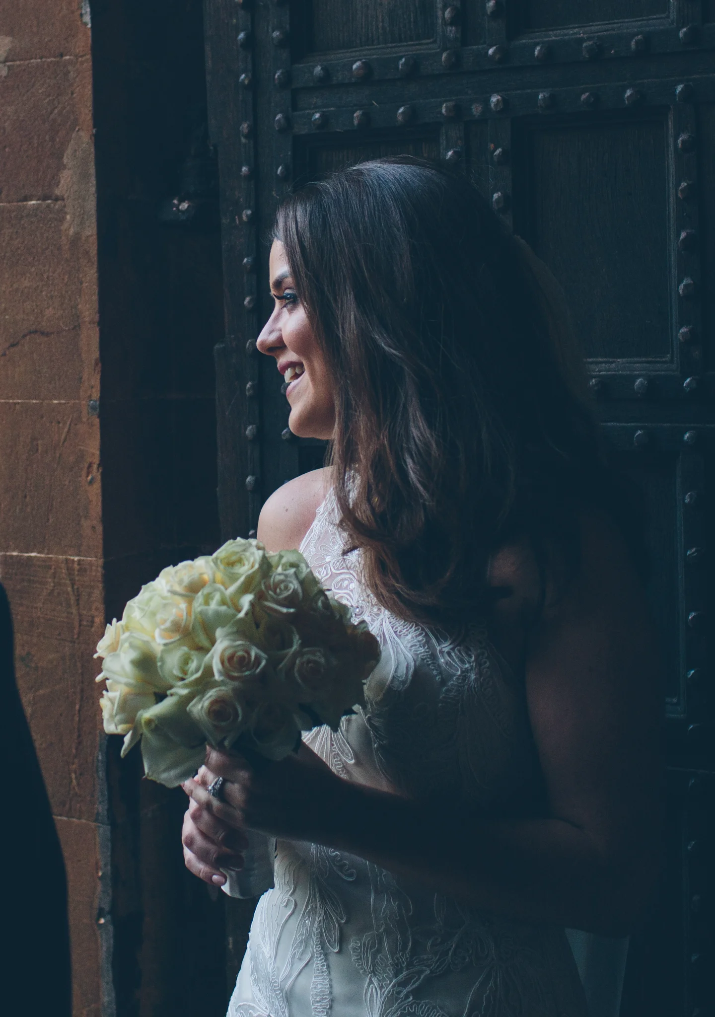 A woman in a white dress is holding a bouquet of white roses, smiling and standing against a dark wooden door.