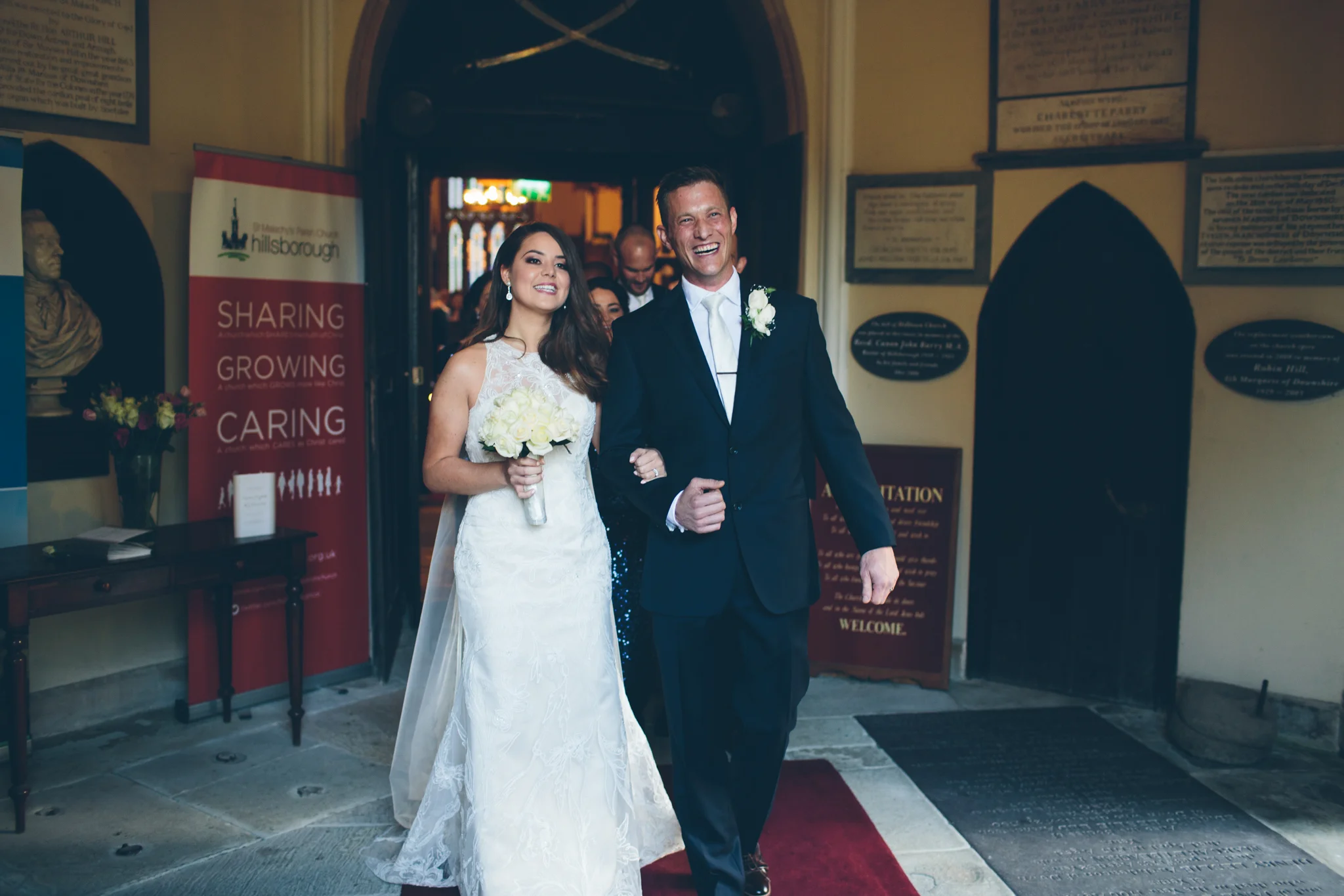 Bride in a white wedding gown holding a bouquet walking arm-in-arm with a groom in a dark suit with a white boutonniere at the entrance of a church, smiling.