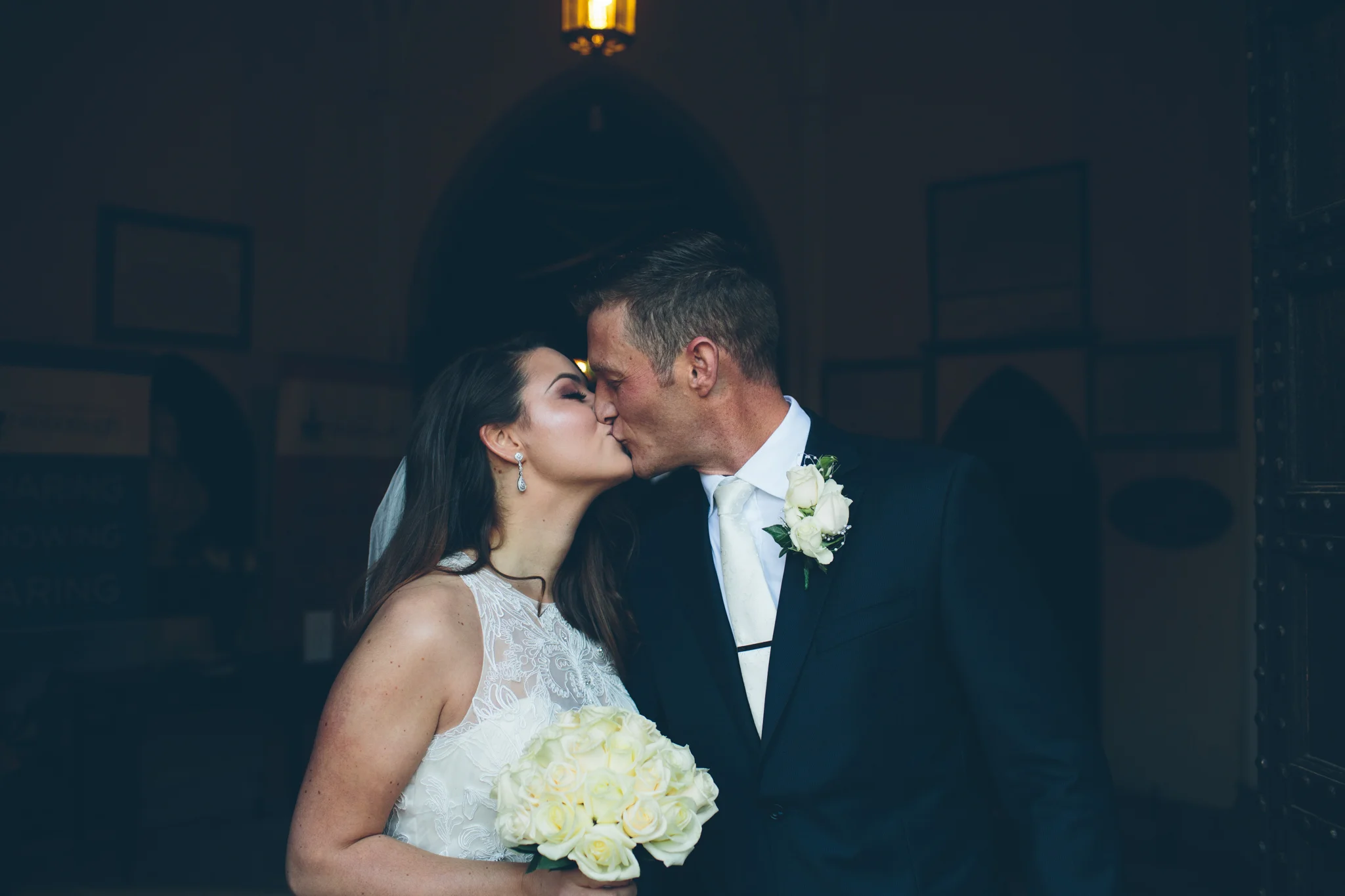 A bride and groom share a kiss indoors at their wedding, with the bride holding a bouquet of white roses and the groom wearing a boutonniere.