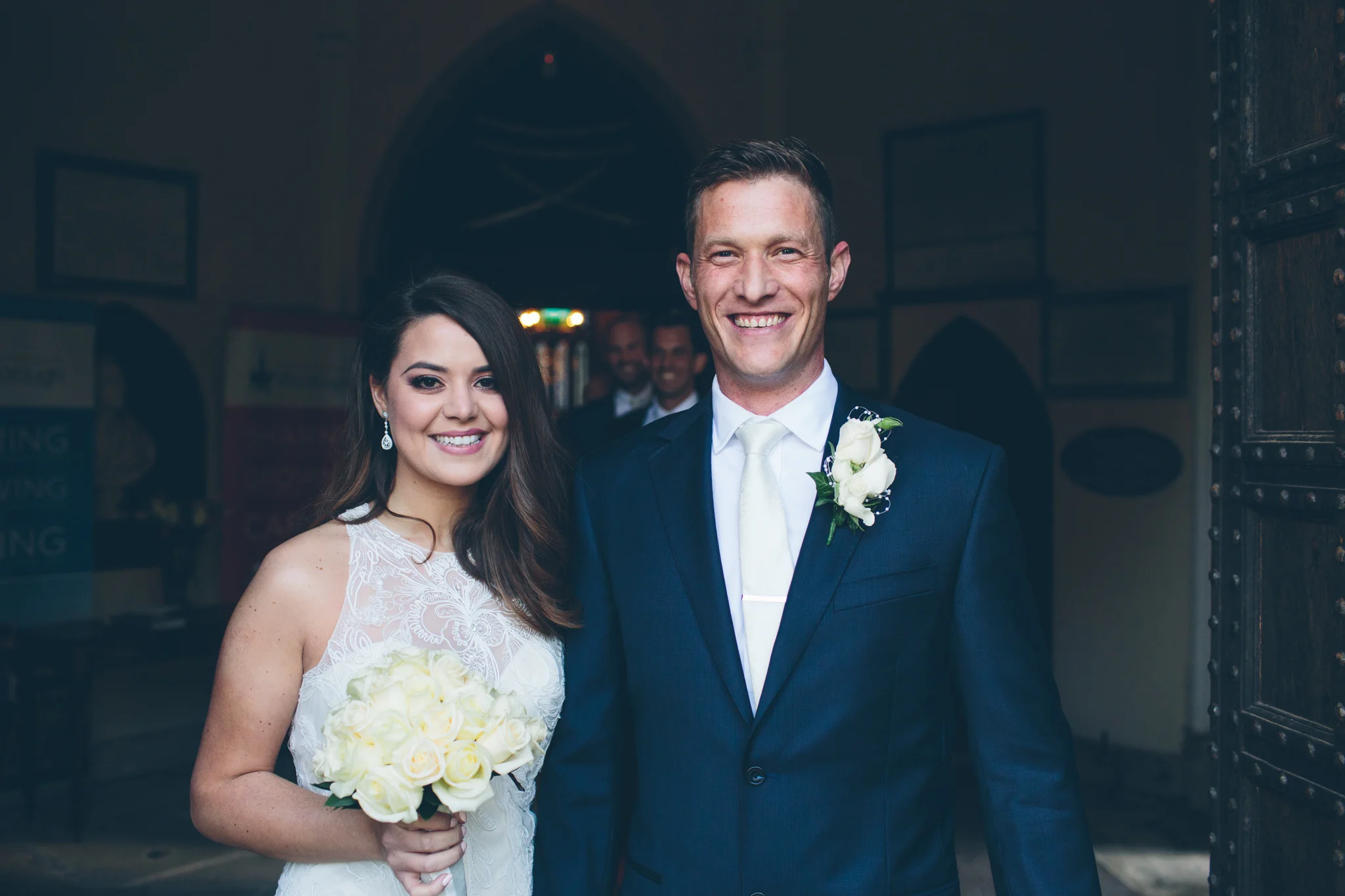 A bride and groom smiling on their wedding day. The bride is holding a bouquet of white roses, wearing a white lace wedding dress with earrings. The groom is dressed in a navy blue suit with a white tie and a white boutonniere.