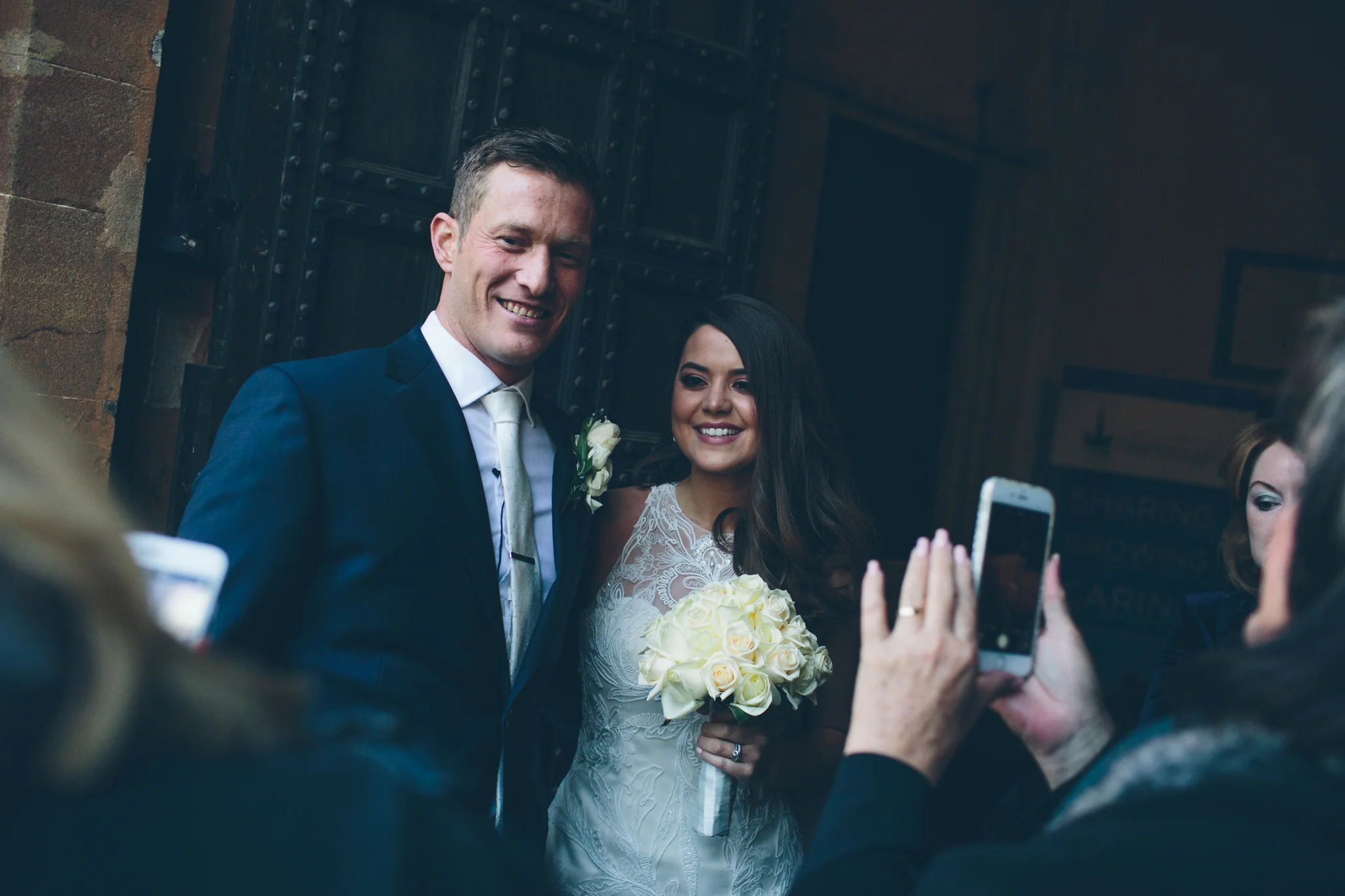 A bride and groom smiling for photos, with the bride holding a bouquet of white roses and the groom in a navy suit and white tie, as guests take pictures in a dimly lit indoor setting.