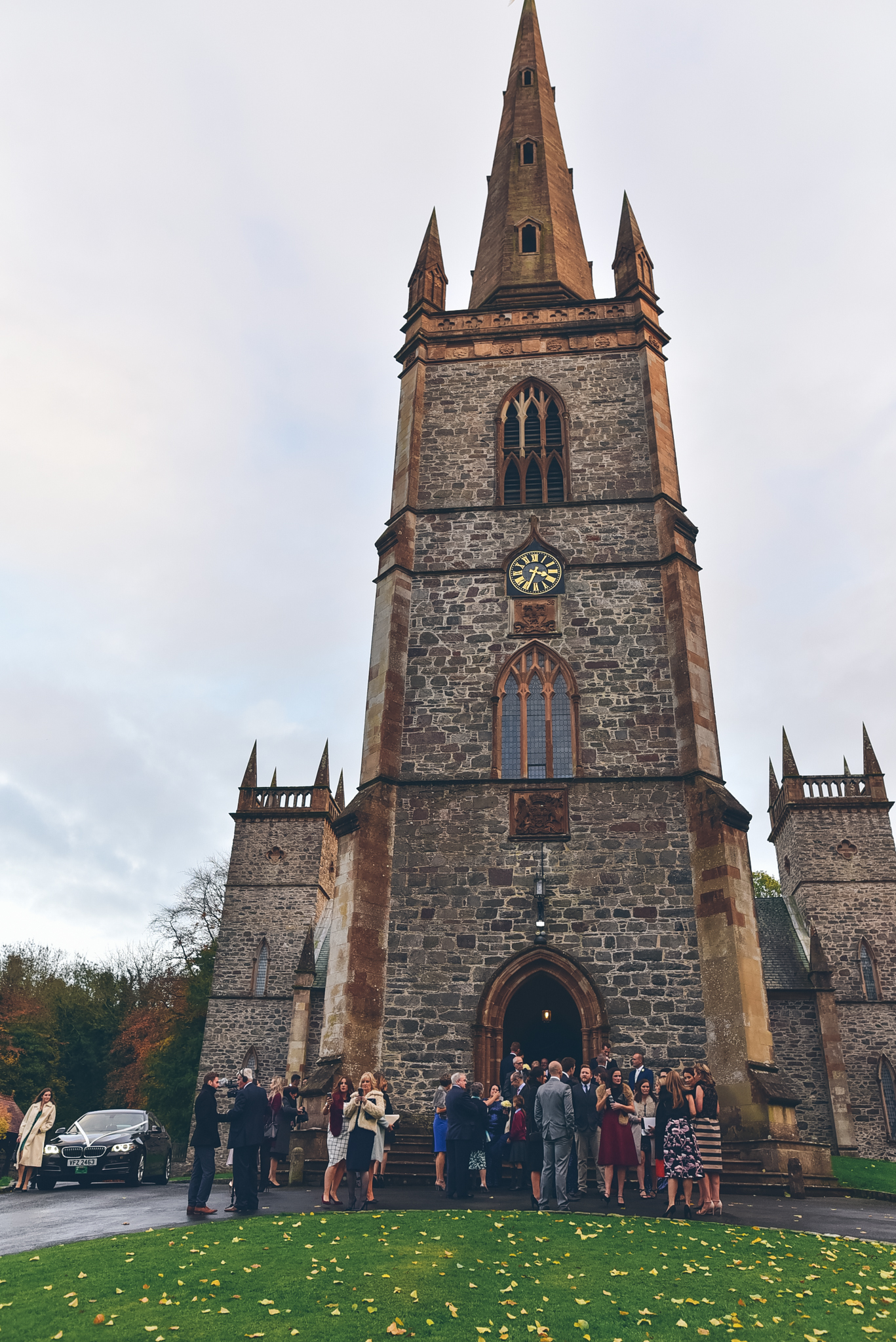 A group of people dressed formally gathering outside a tall stone Gothic-style church with a clock on its tower, during an overcast day.