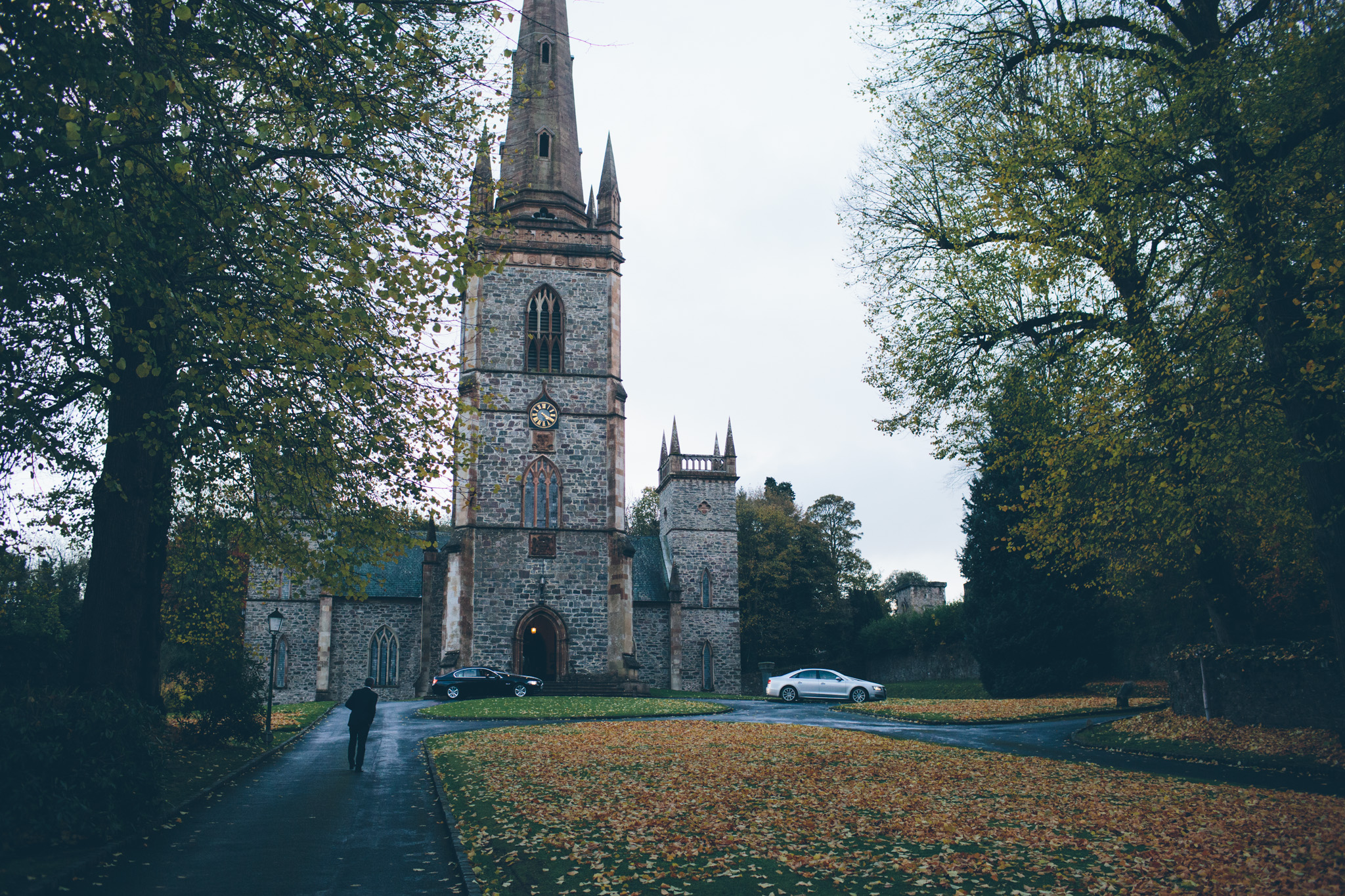 A stone church with tall spires and a large clock on the front tower, surrounded by trees and fallen autumn leaves, with a person walking on a paved path.