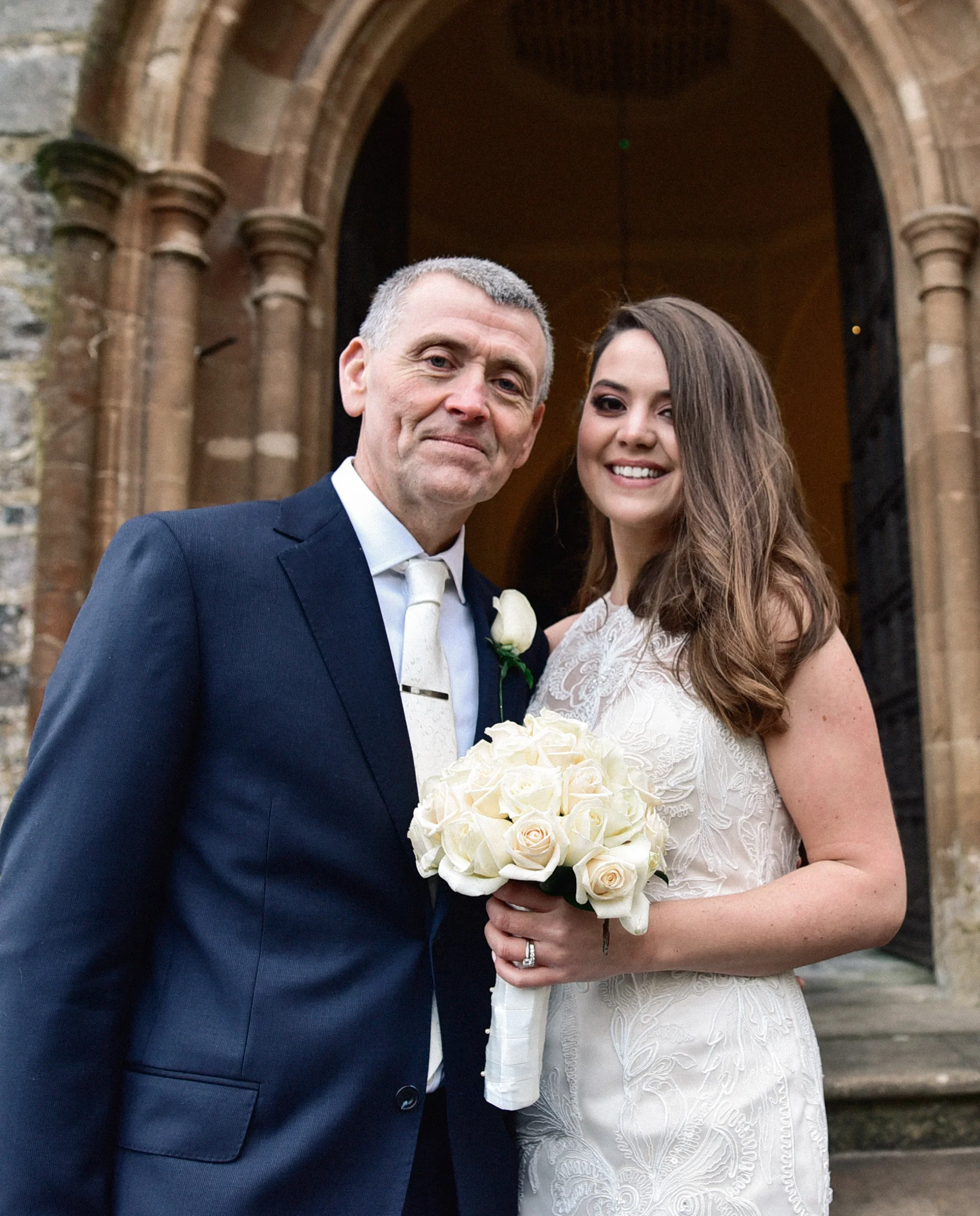 A father and daughter at her wedding, standing outside a stone church entrance. The father is wearing a navy suit and white tie, smiling. The bride has long brown hair, wearing a white lace wedding dress and holding a bouquet of white roses, smiling.