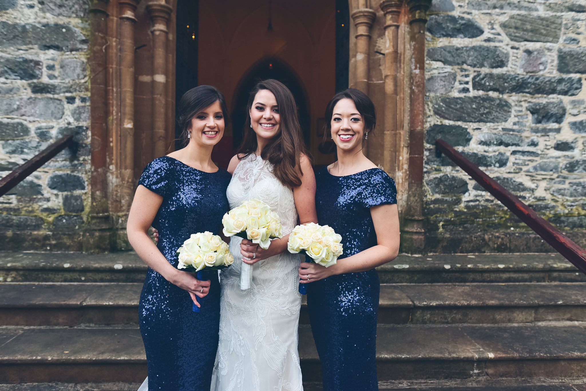 Three women, one bride in a white wedding dress holding a bouquet of white roses, flanked by two bridesmaids in matching dark blue sequin dresses each holding a bouquet of white roses, standing on stone steps outside a historic stone church with wood