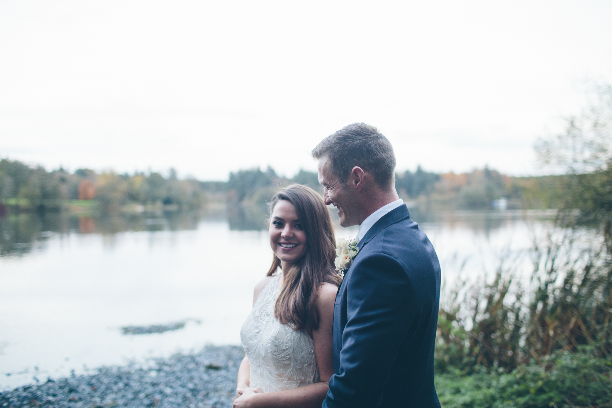 A newlywed couple standing near a lake, smiling and enjoying each other's company during daytime.