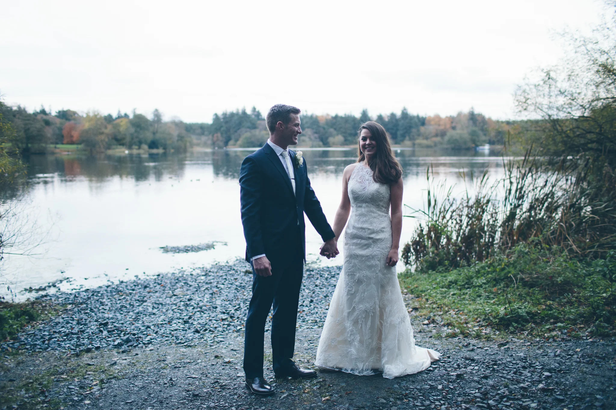 A newlywed couple holding hands by a lakeside during daytime, with trees and water in the background.