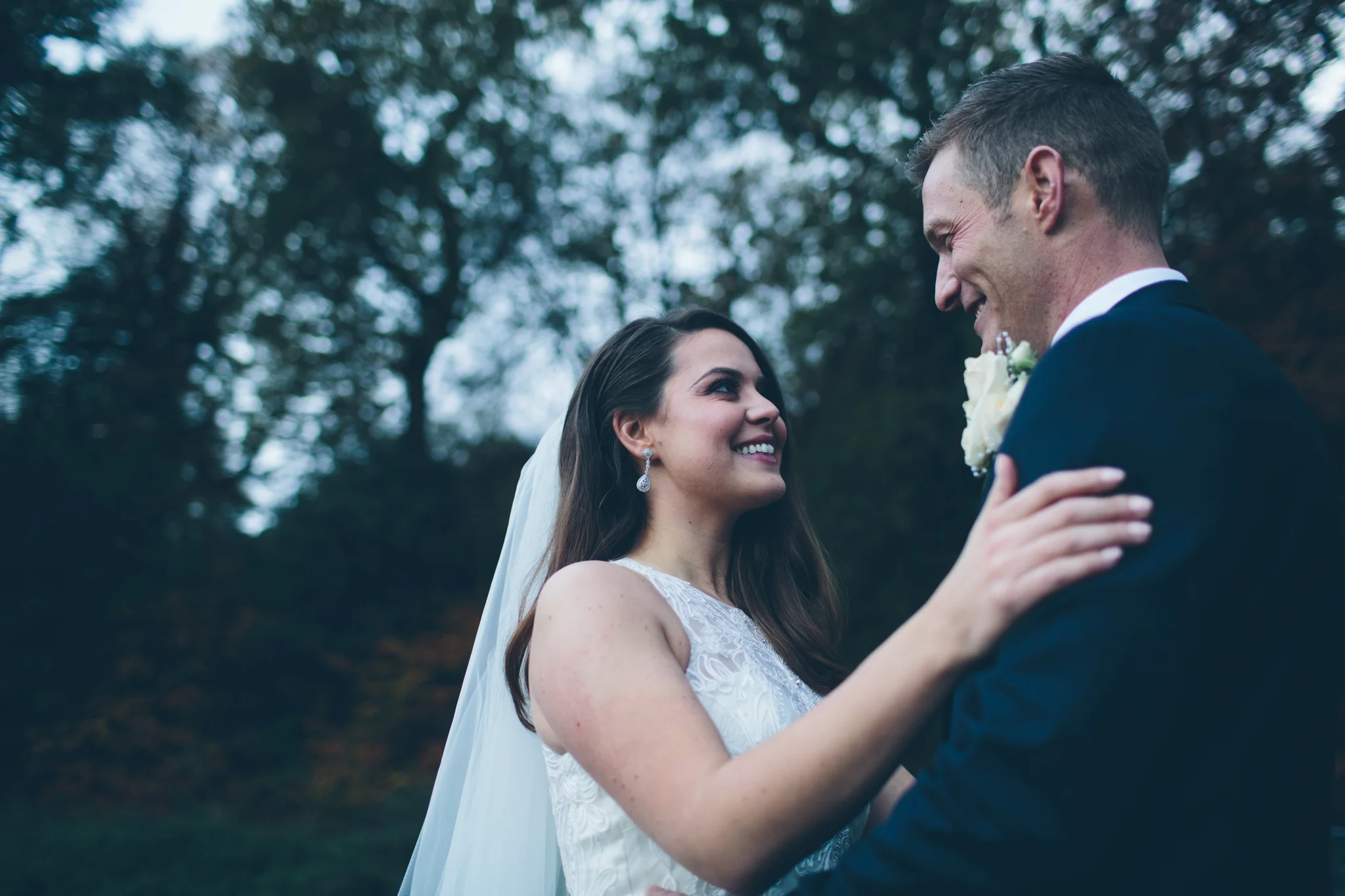 A bride and groom share a kiss outdoors with trees in the background, smiling at each other.
