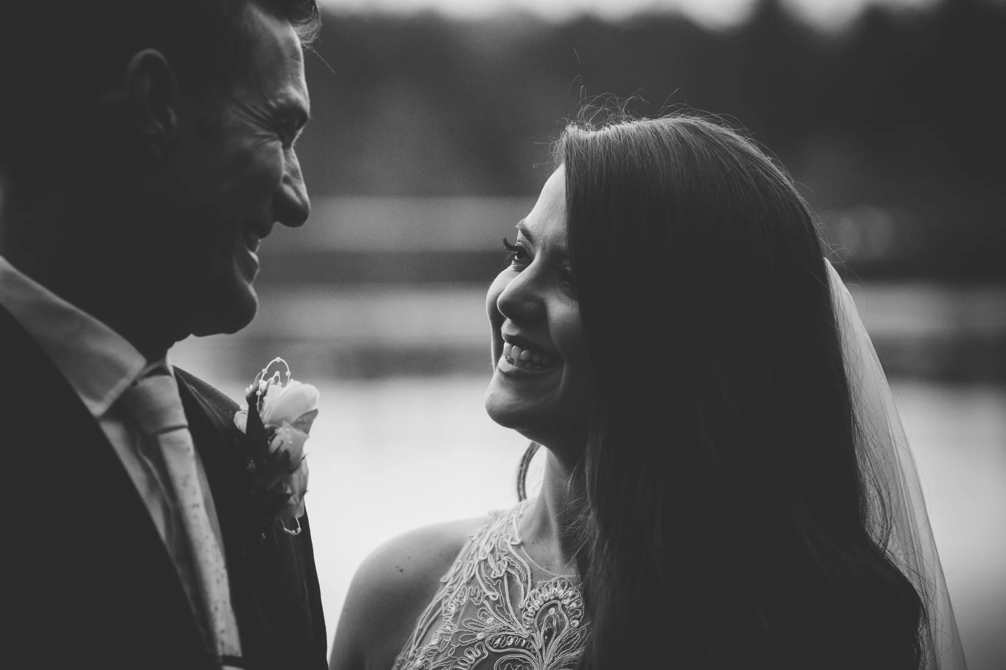A black and white photo of a happy couple smiling at each other outdoors, with a blurred background.
