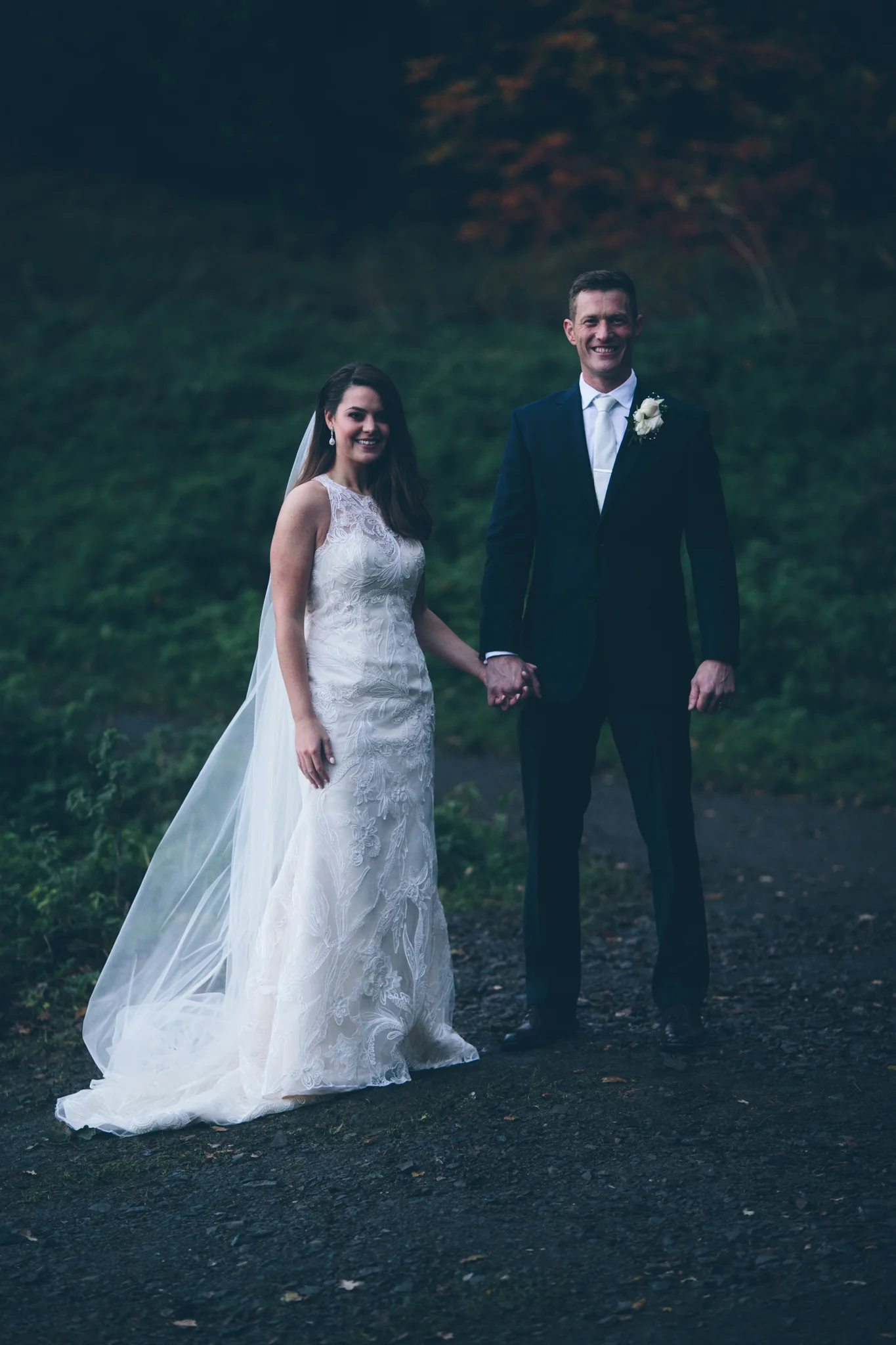 A bride and groom holding hands outdoors during their wedding, standing on a dark path with greenery and trees in the background.