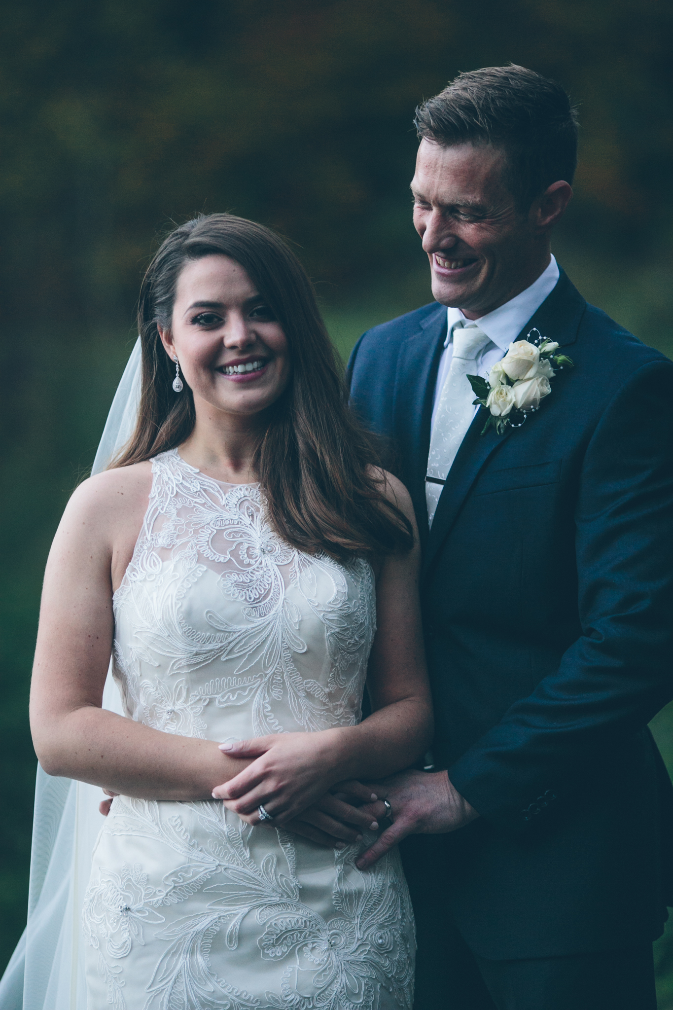 A bride and groom smiling at their wedding photos outdoors, with the groom touching the bride's waist, both dressed in wedding attire.