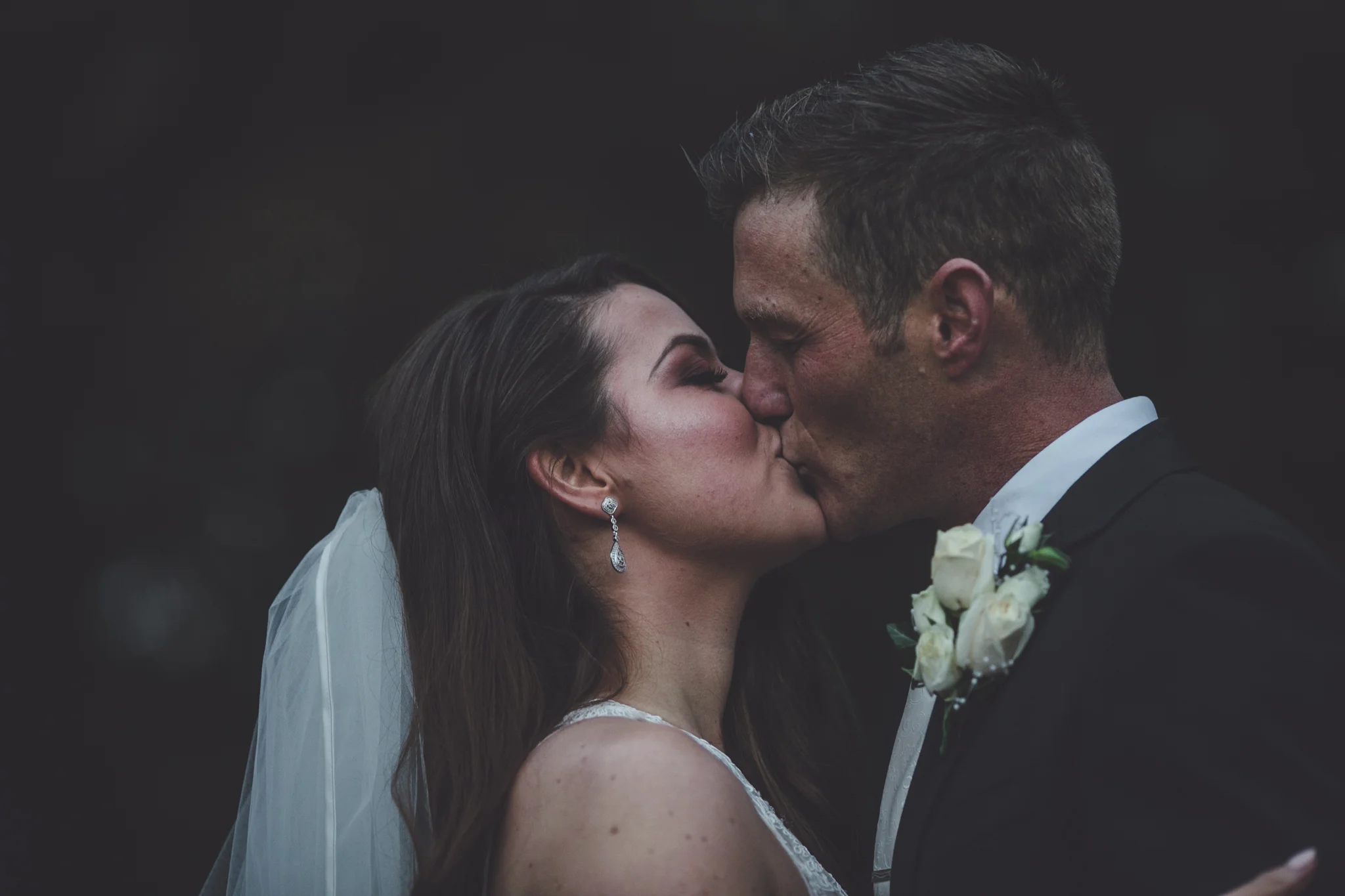 A bride and groom kissing during their wedding ceremony, with the bride wearing a veil and earrings, and the groom wearing a suit with a white boutonniere.
